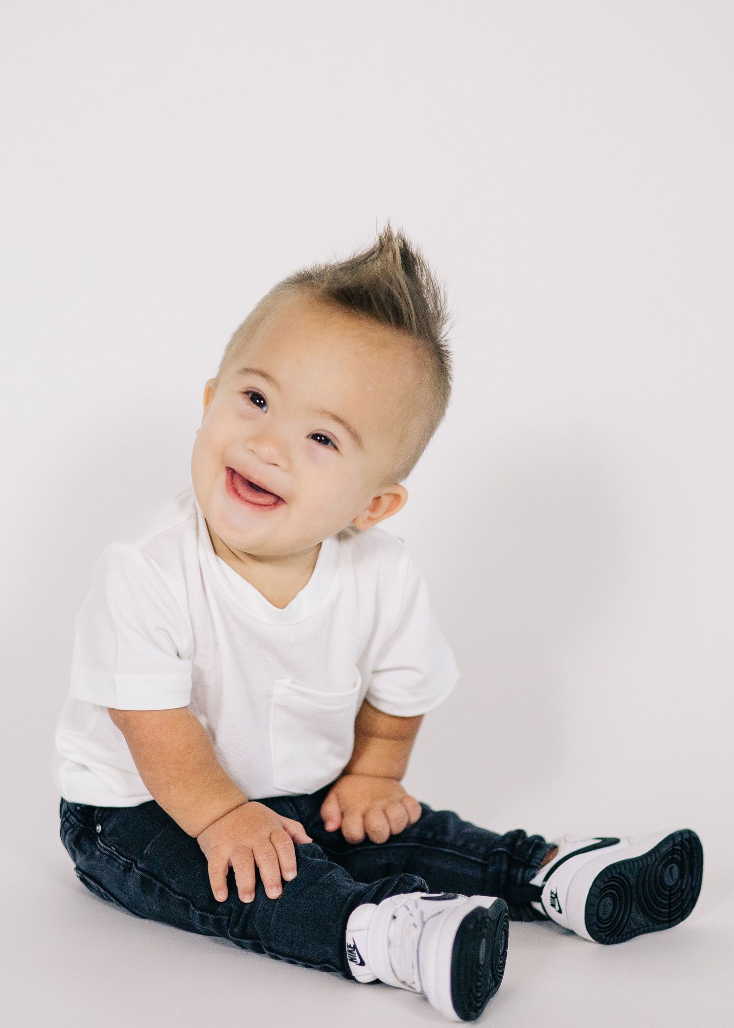 Baby in white t-shirt and dark pants sitting on white background.