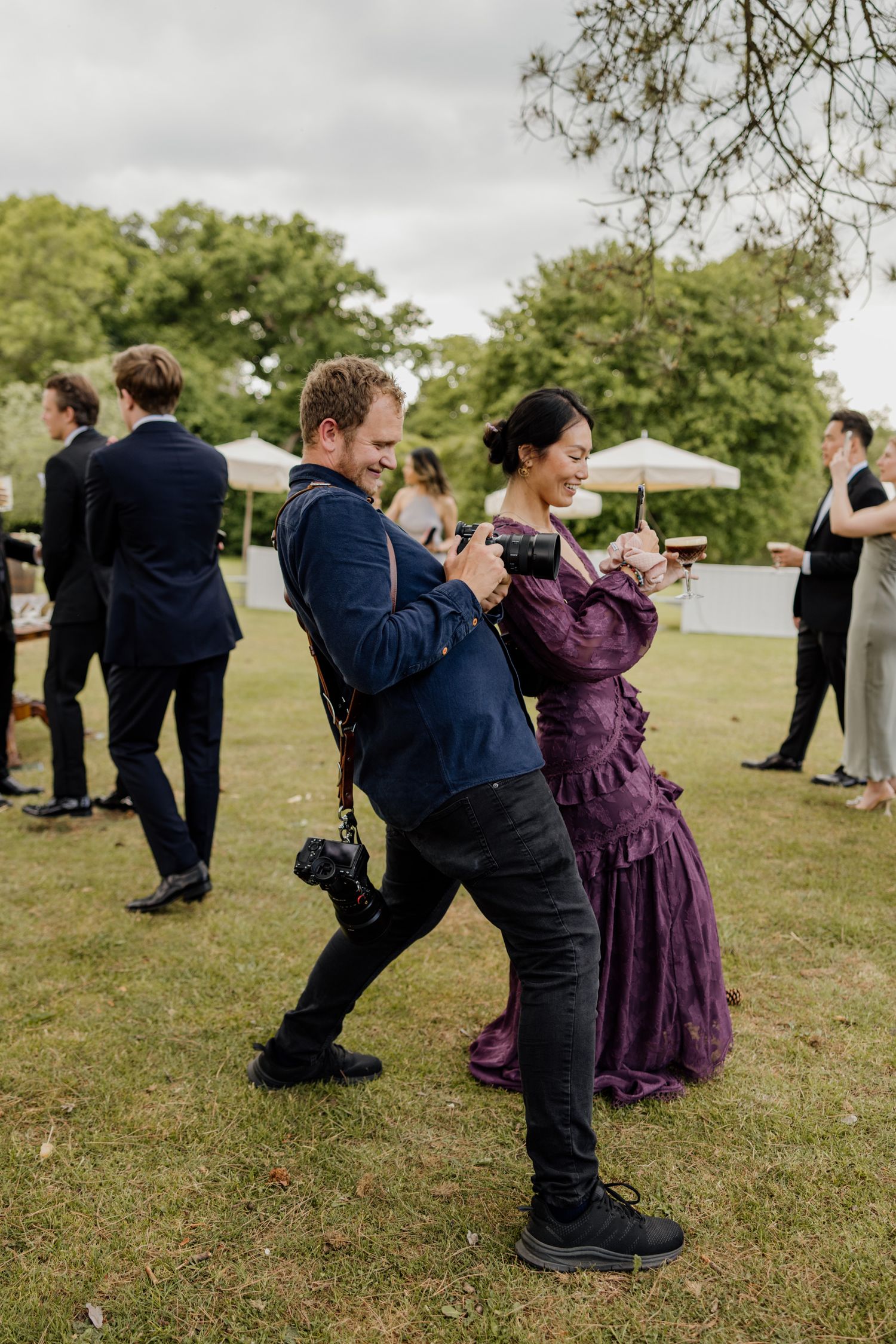 A man in a navy suit and woman in a purple Victorian dress laugh and dance together at an outdoor garden wedding reception.