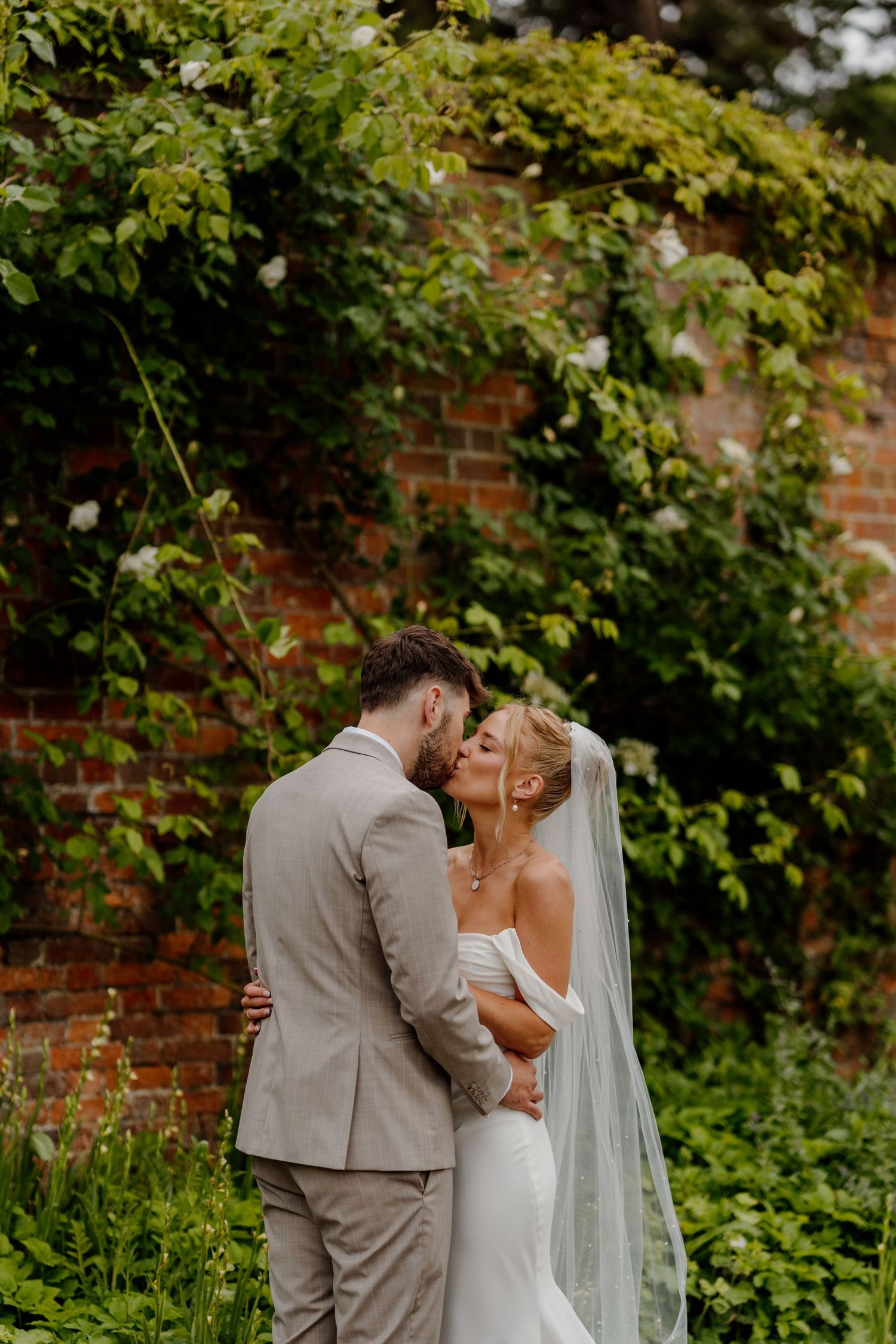 A bride and groom share a tender kiss in a lush garden beside a rustic brick wall, the bride wearing a white gown and veil.