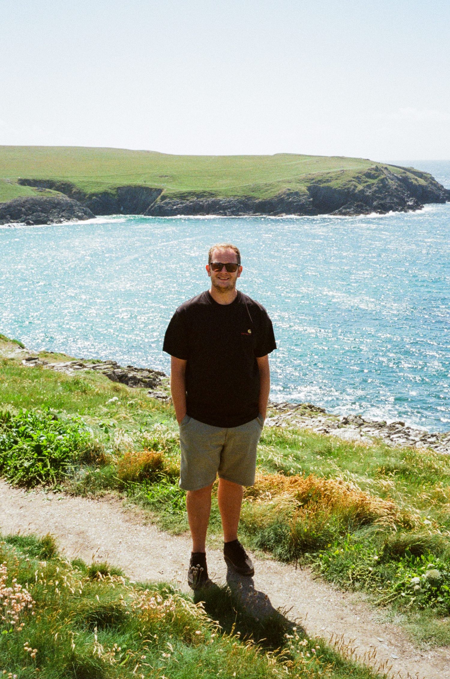 Man in black t-shirt and shorts standing on coastal path with sparkling blue sea and green cliffs behind him.