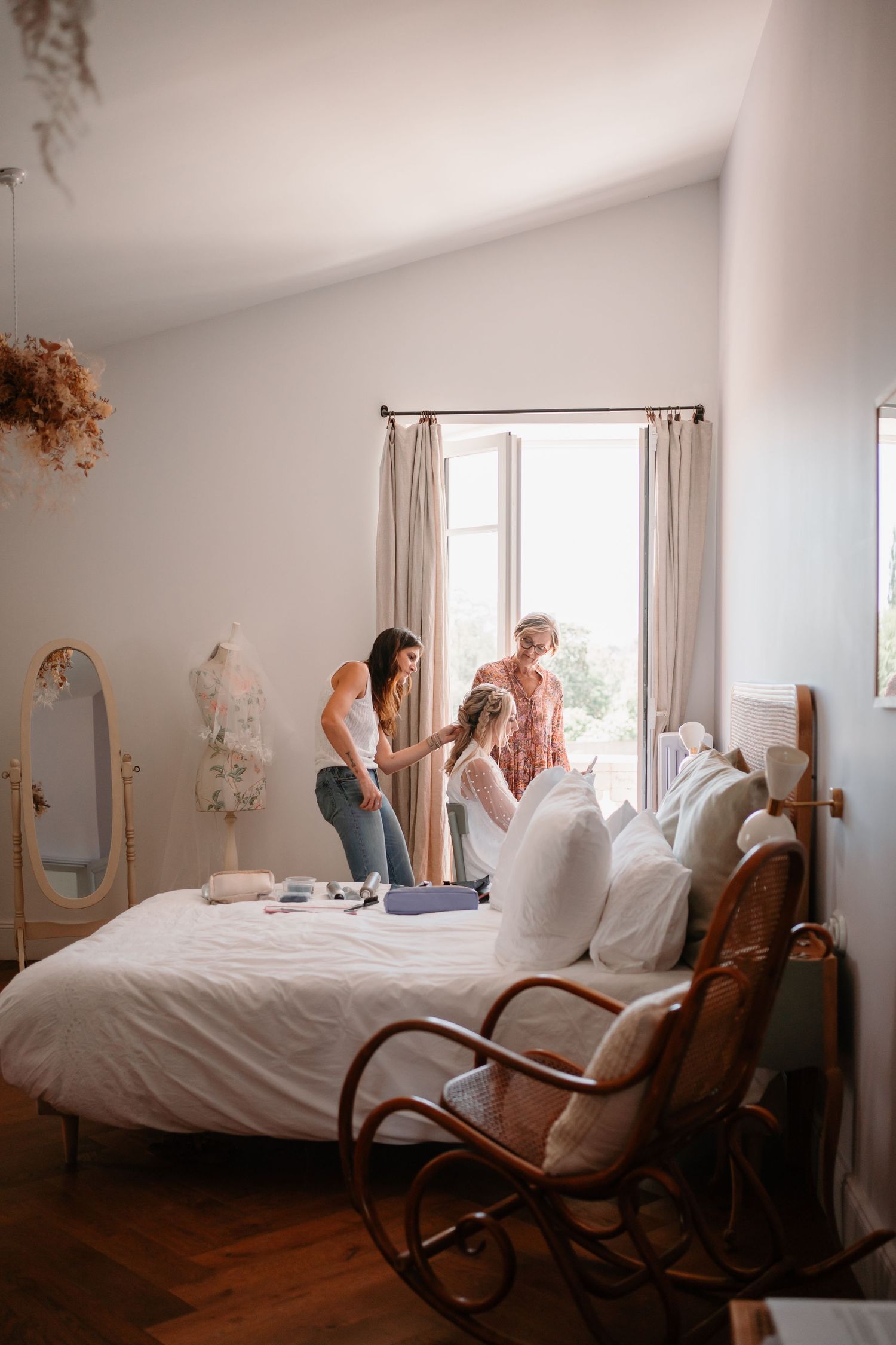 A cozy bedroom with white bedding, vintage rocking chair, and natural light streaming through curtained windows.