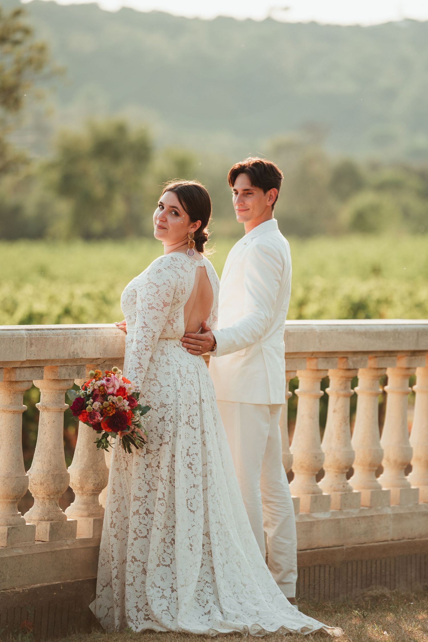 A couple in white wedding attire share an intimate moment on a stone balcony overlooking a scenic landscape at sunset.