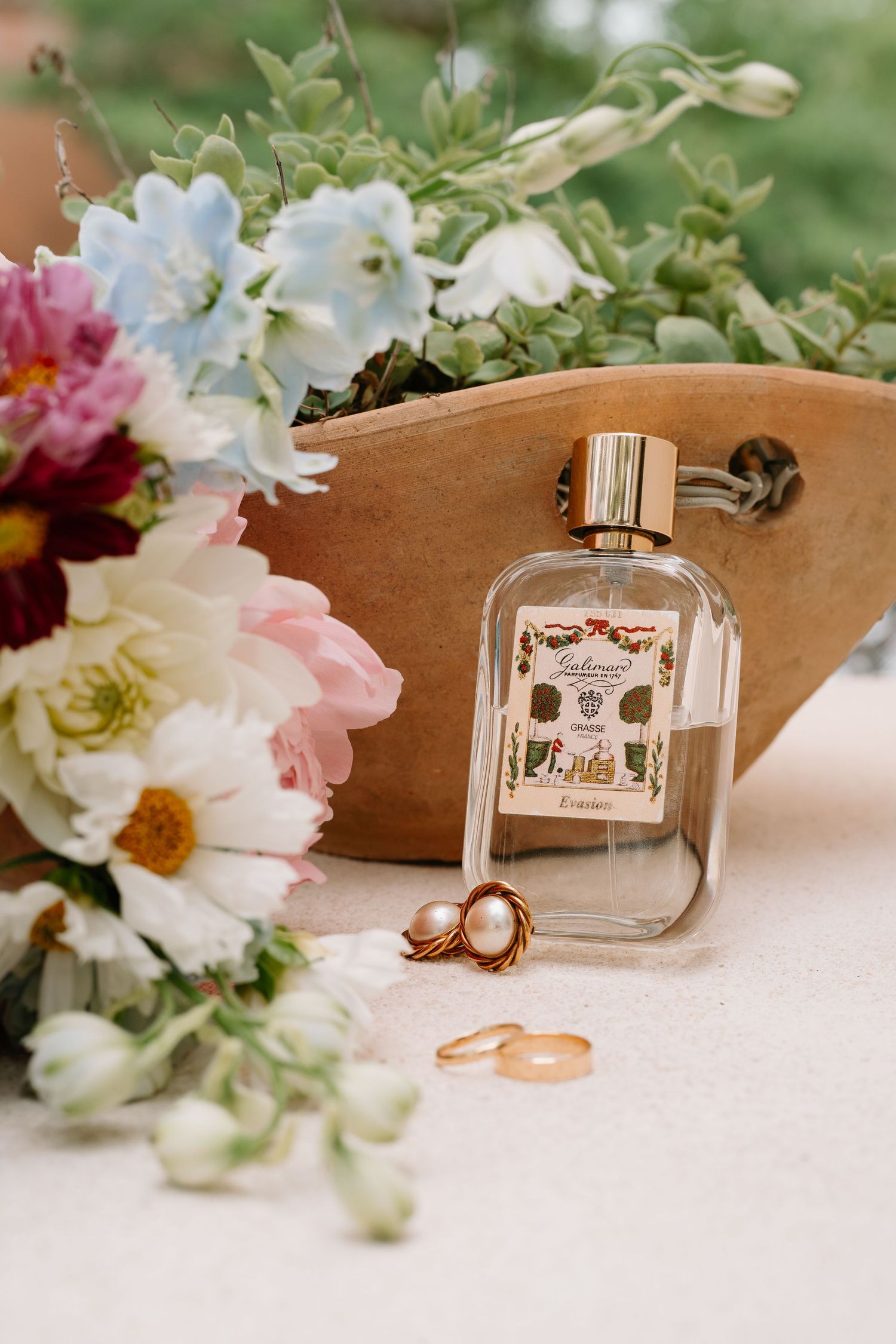 Elegant wedding flat lay with perfume bottle, gold rings, pearl earring, and colorful floral bouquet on soft linen.
