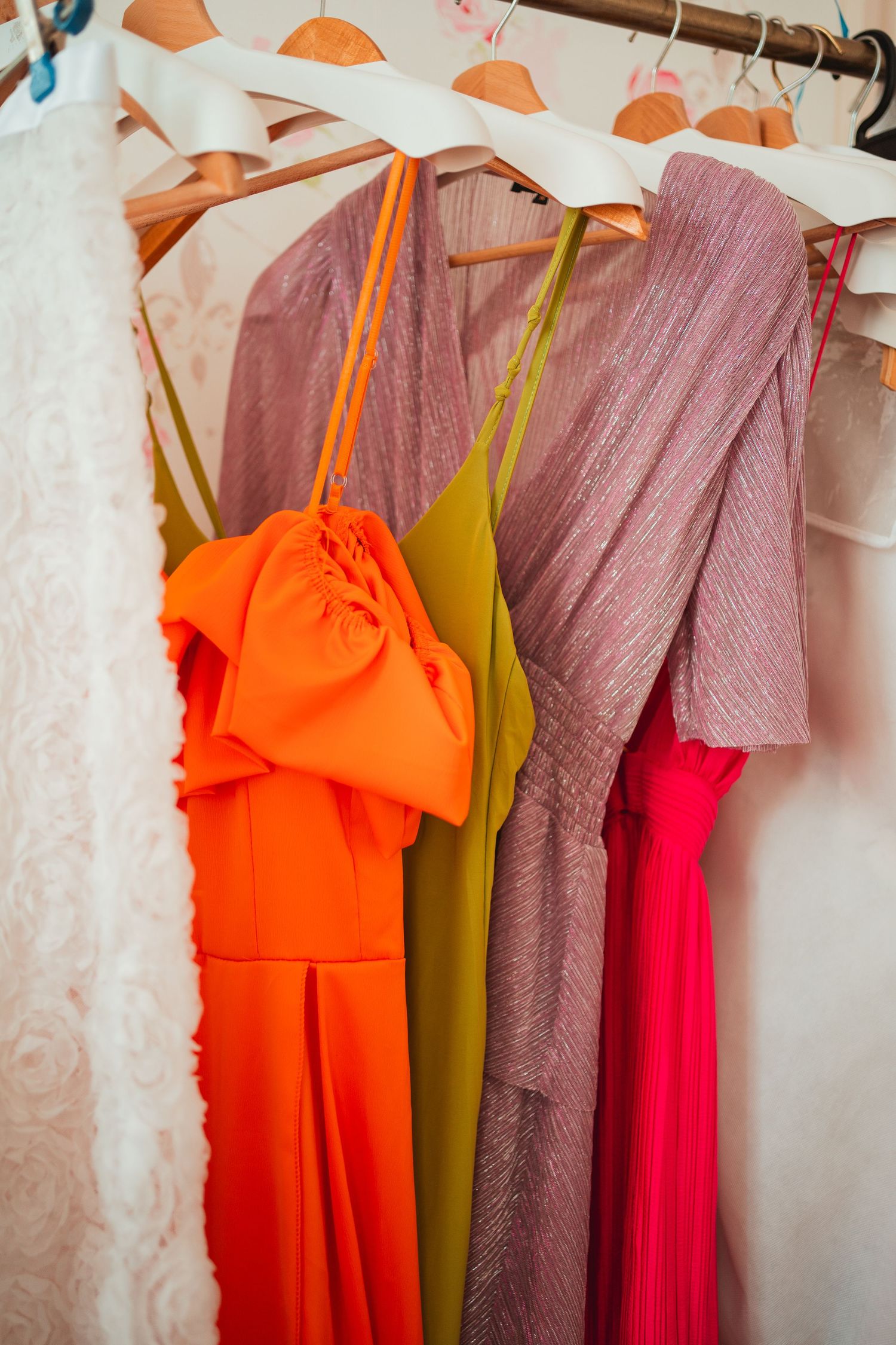 Colorful bridesmaid dresses including orange, olive green, and pink hanging on a white curtained wall.