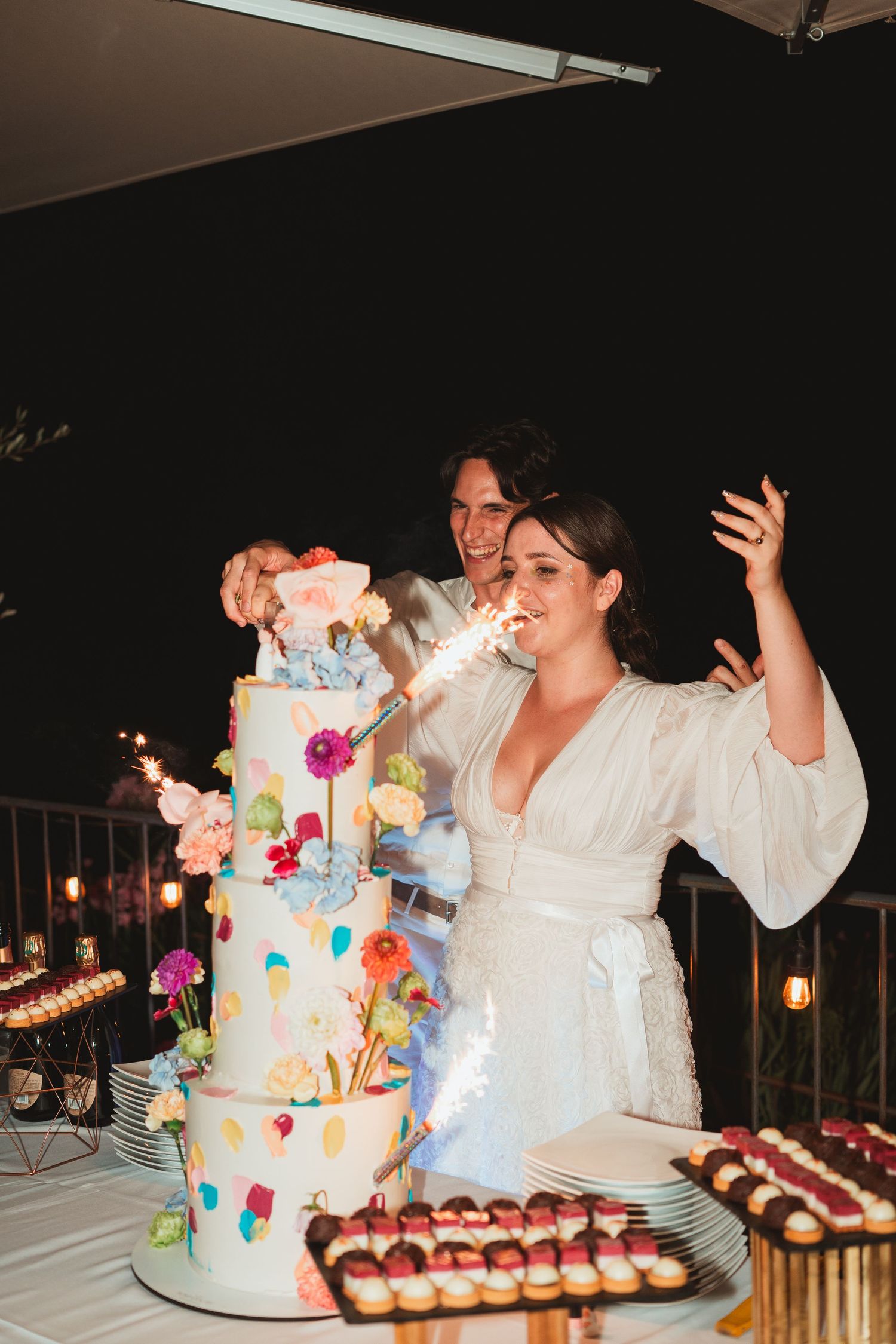 A person in white reacts with excitement while cutting into a colorful multi-tiered wedding cake at a reception.