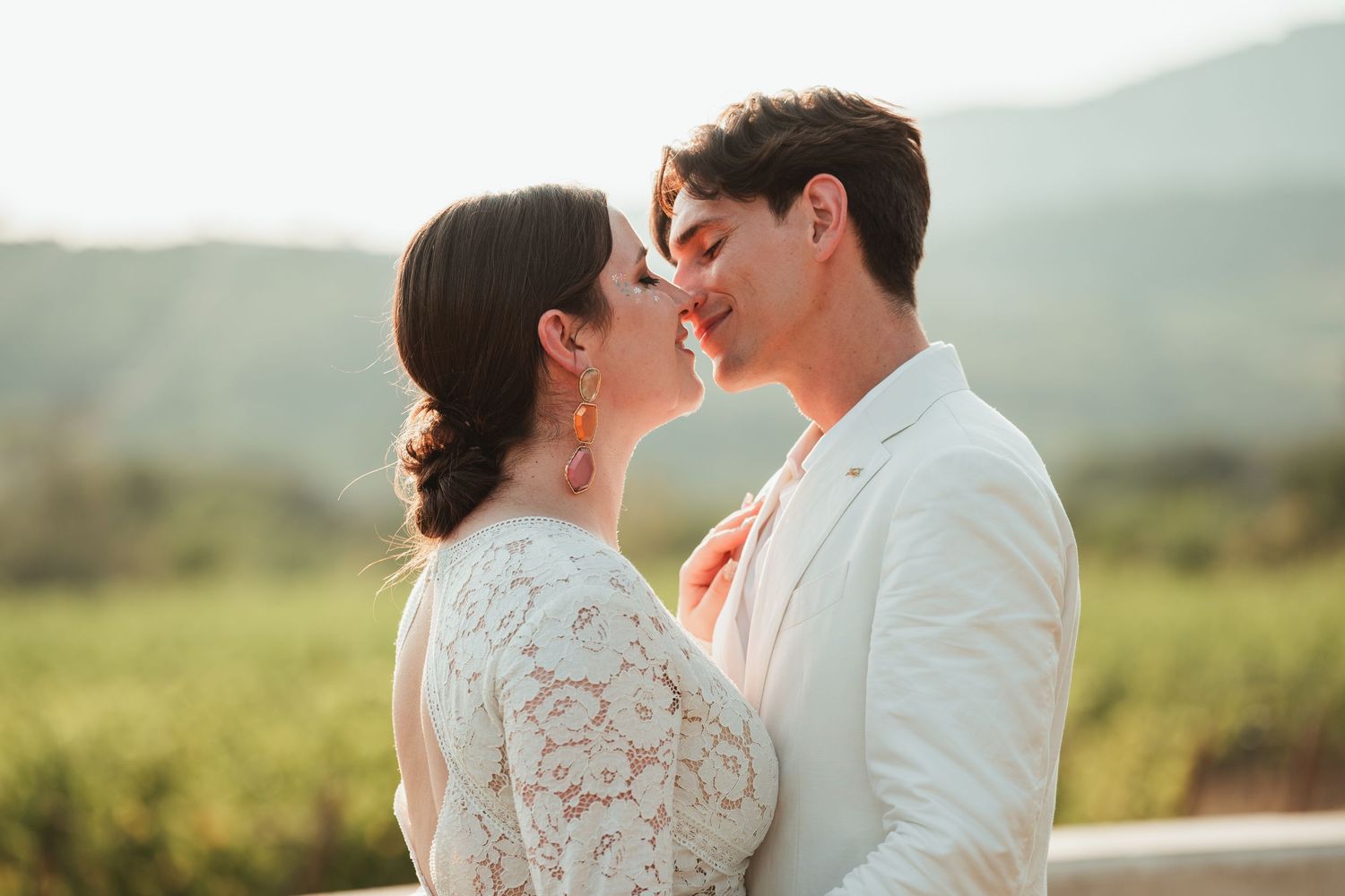 A couple in wedding attire share an intimate moment in a scenic outdoor setting with rolling hills in the background.