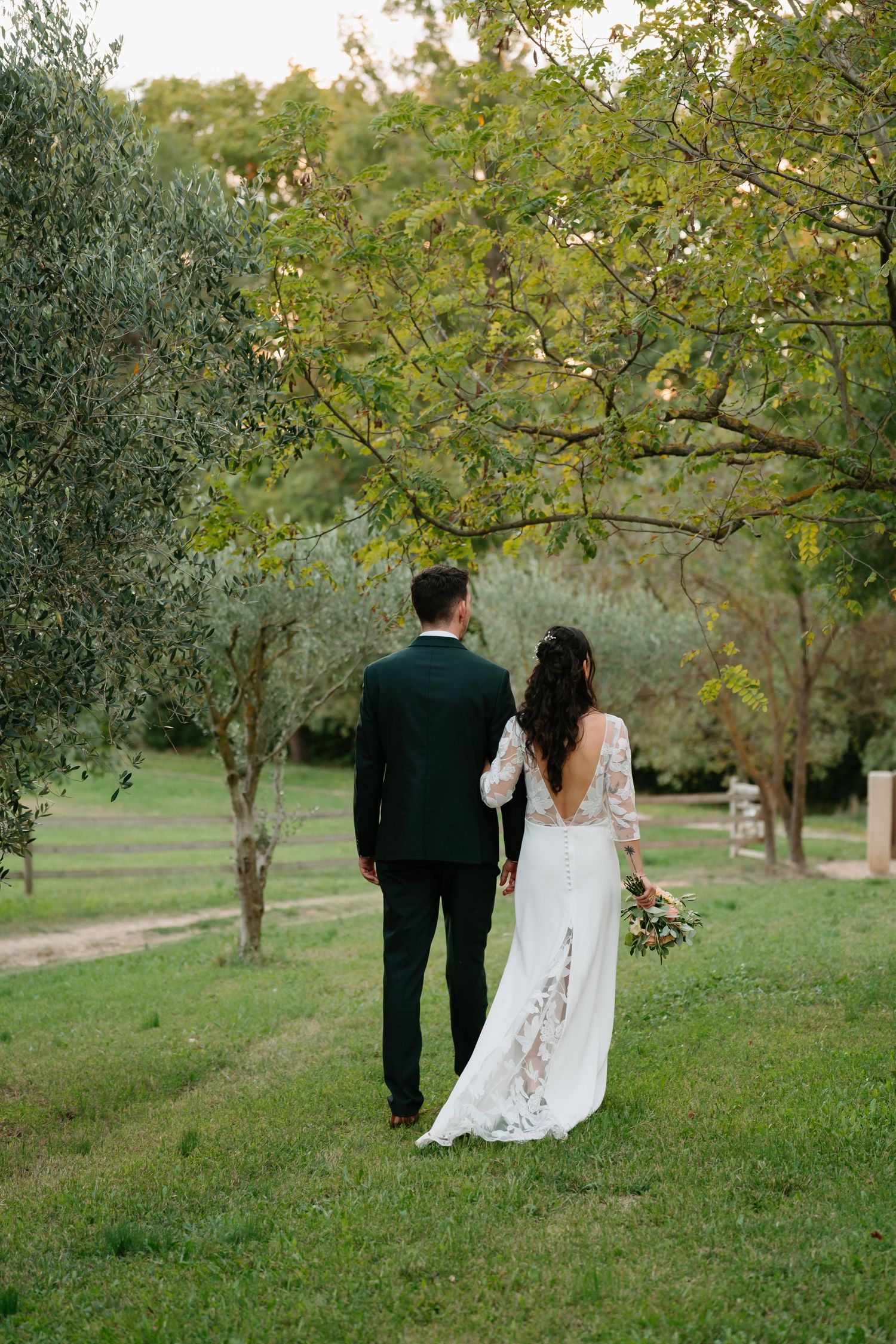 A couple in wedding attire walk together through a lush garden setting with trees and greenery.