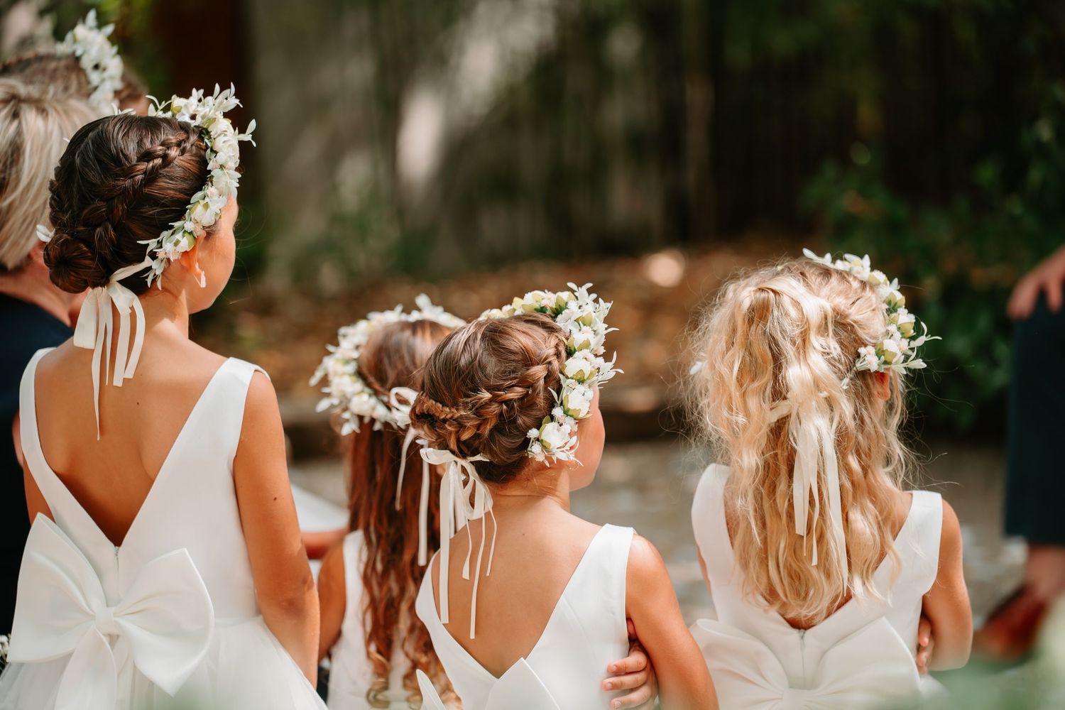 Row of bridesmaids in white dresses with floral crowns stand together during outdoor wedding ceremony.