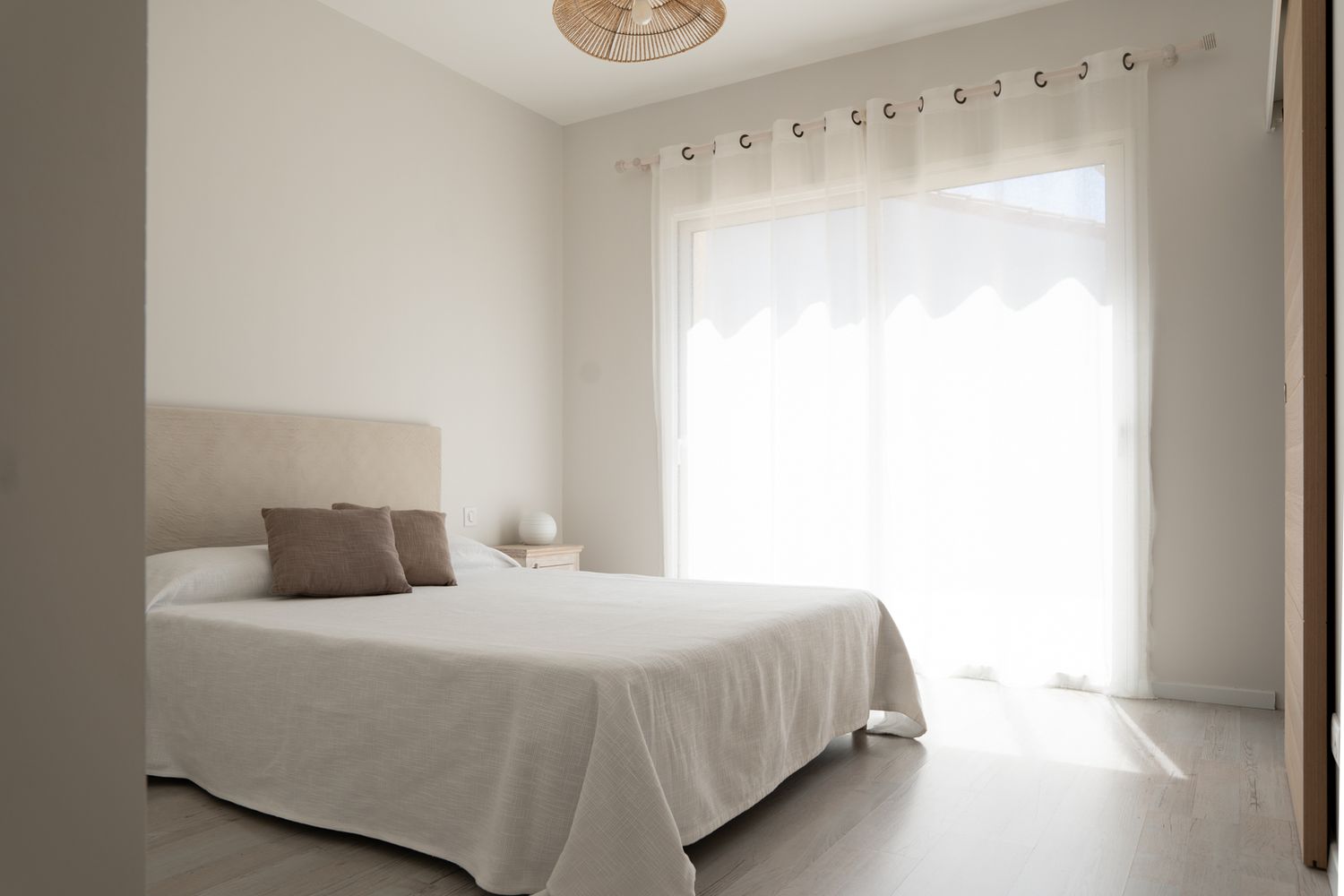 A bright, minimalist bedroom interior with white bedding, natural light streaming through windows, and neutral wall tones.