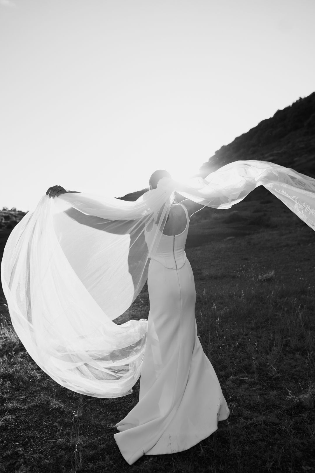 Black and white photo of a bride in a flowing white gown, her veil billowing in the wind on a scenic hillside.