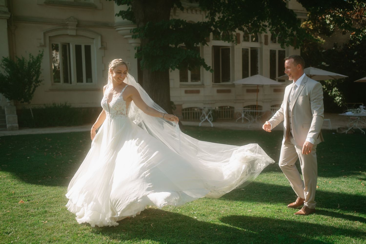 A flowing white wedding dress billows in the breeze as a couple walks through a sunlit garden setting.