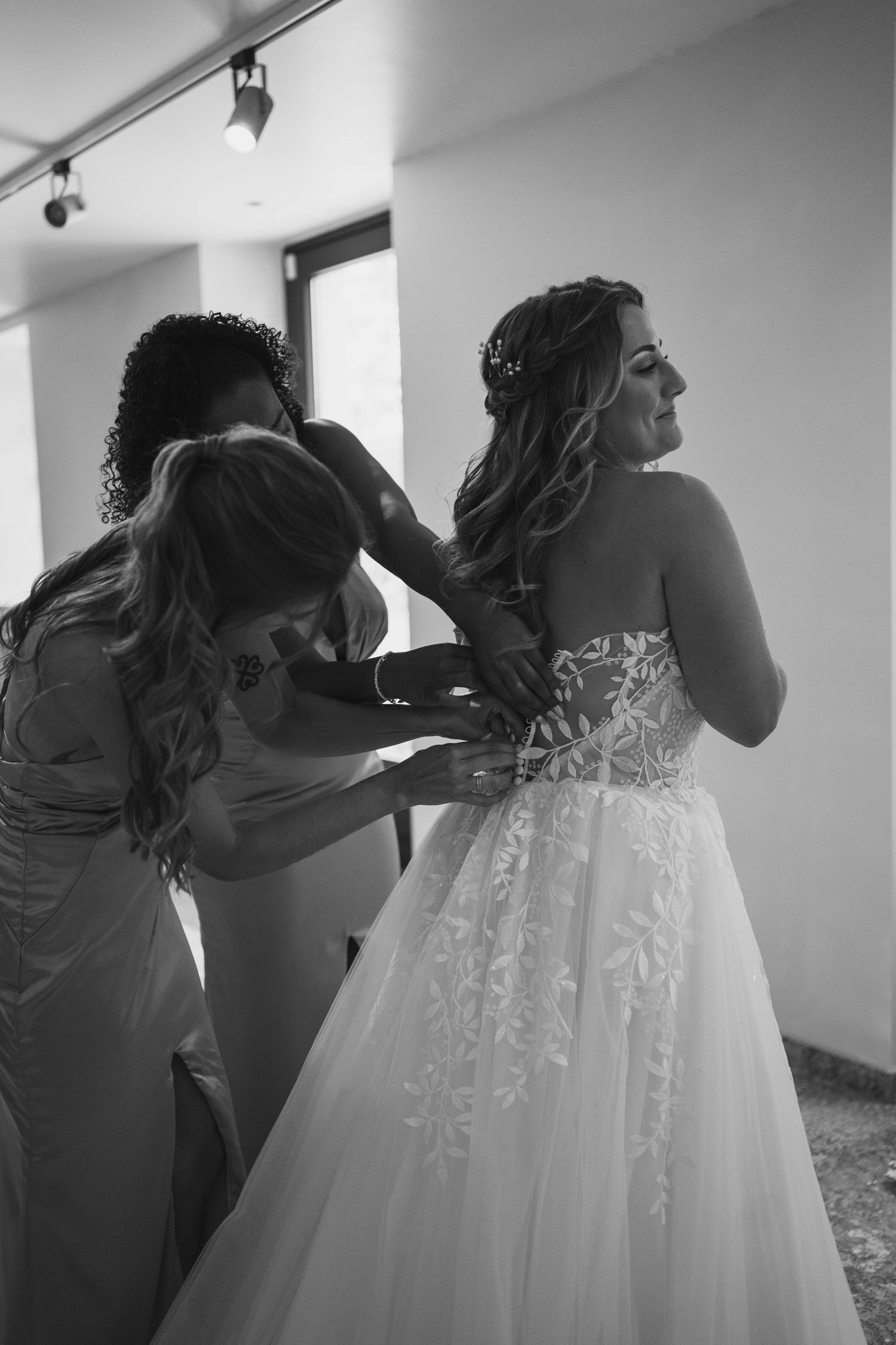 A black and white photo showing final adjustments being made to a bride's elegant lace wedding gown.