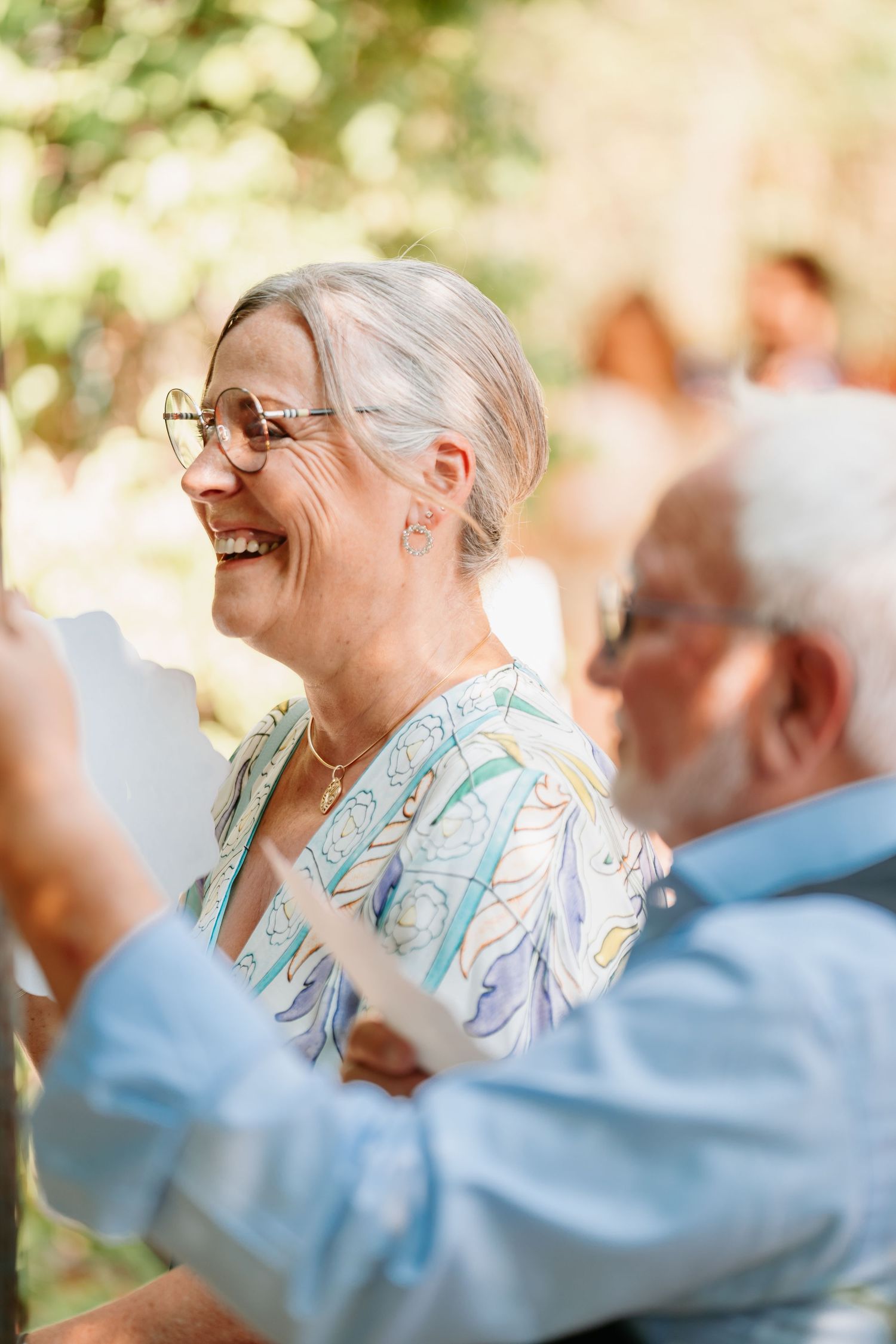 Joyful senior in glasses and floral print dress laughing outdoors with others during a sunny gathering.
