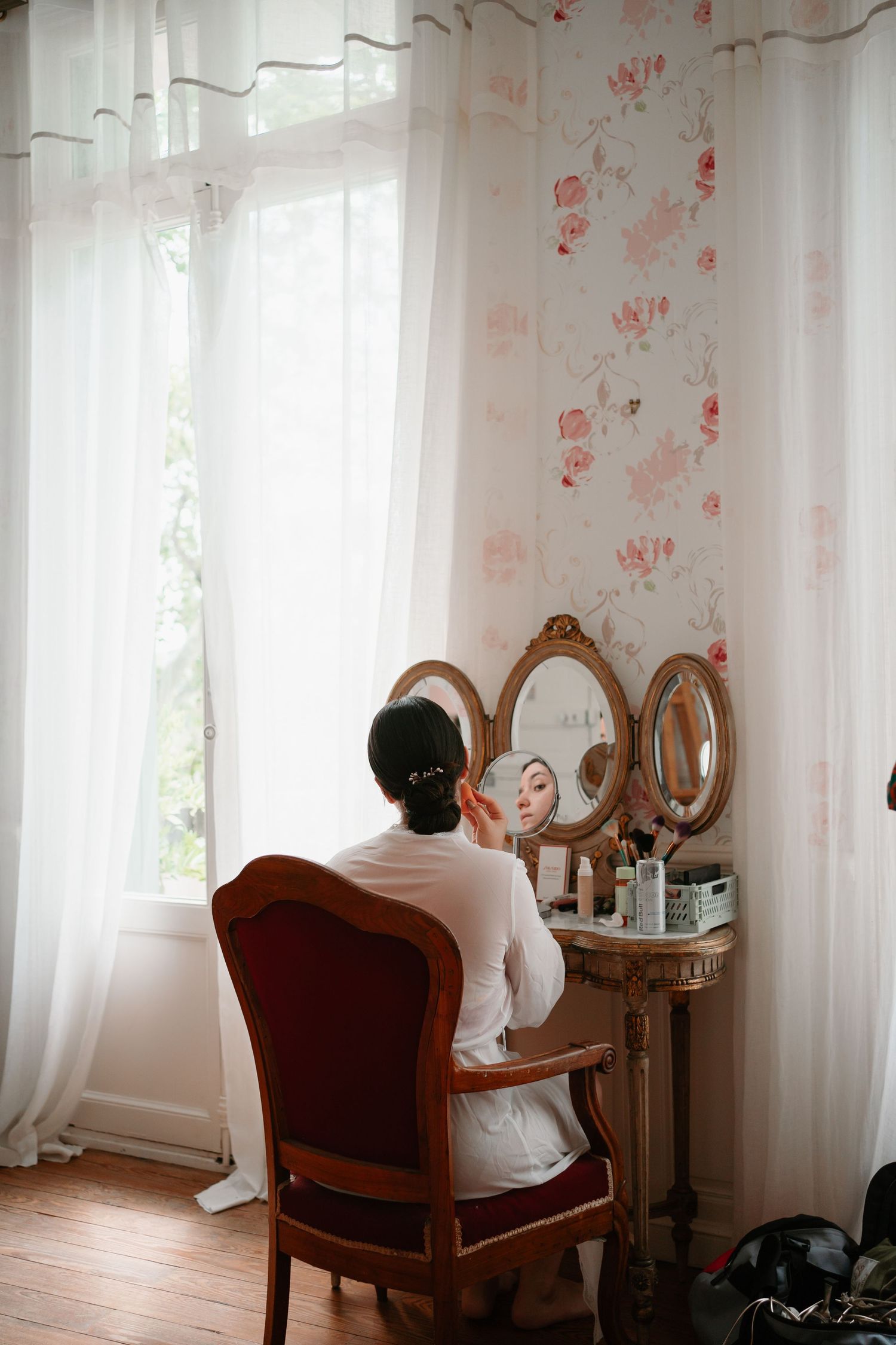 A vintage vanity table with triple mirrors sits by a window with sheer curtains and floral wallpaper in a bright room.