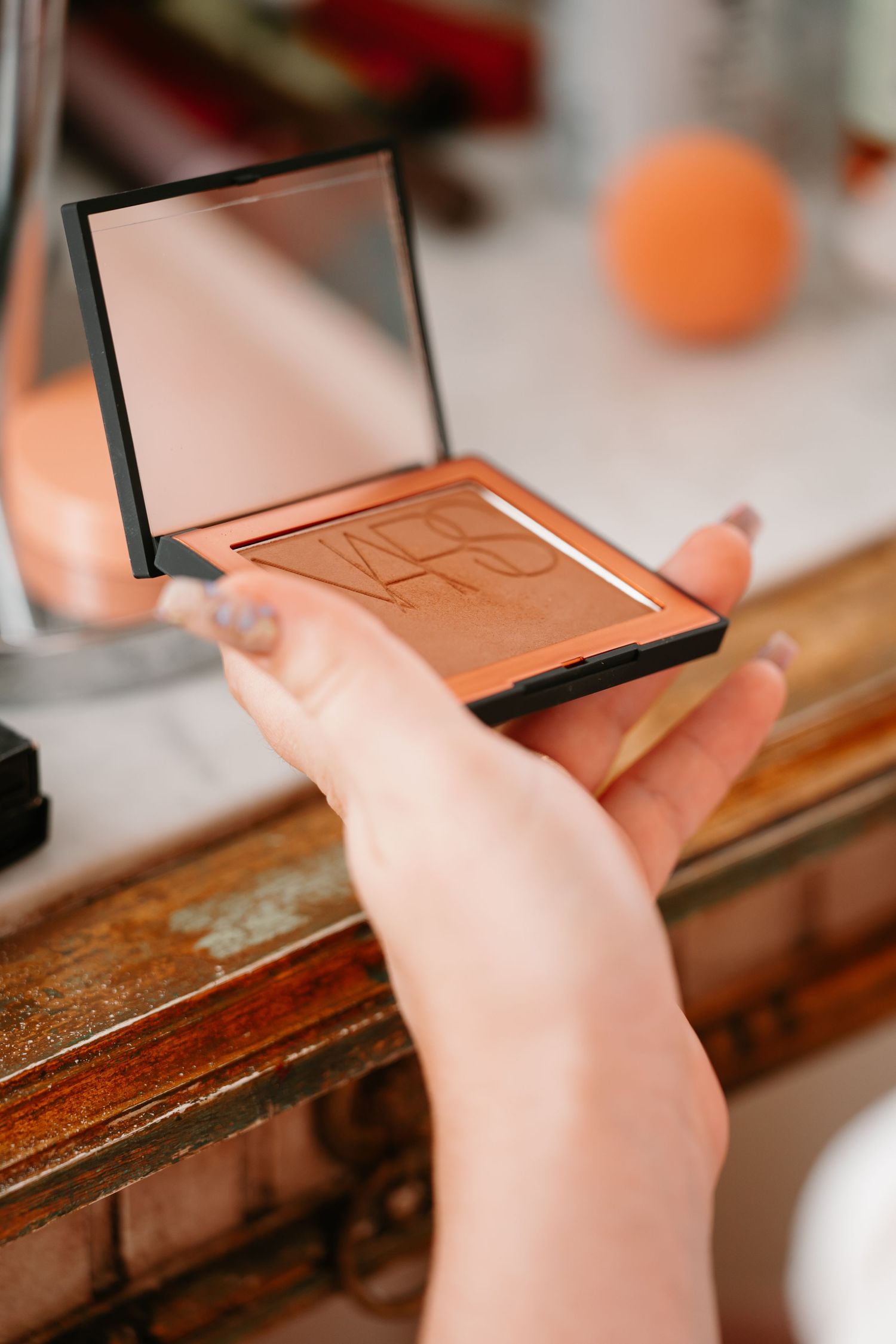 Hands holding an open bronzer or face powder compact makeup palette with mirror on a wooden surface.