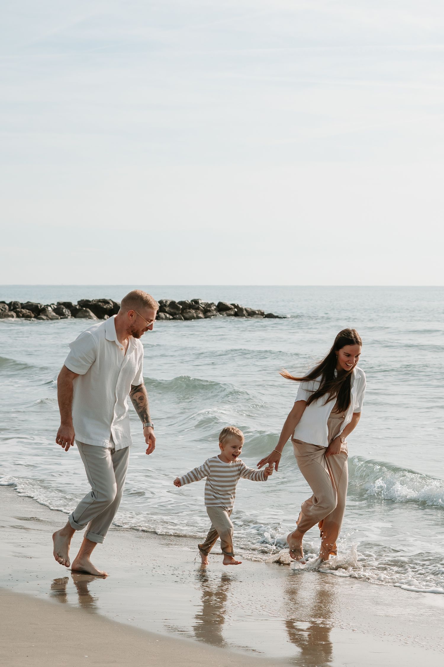 A family walks together holding hands along a beach with waves and a rocky jetty in the background.