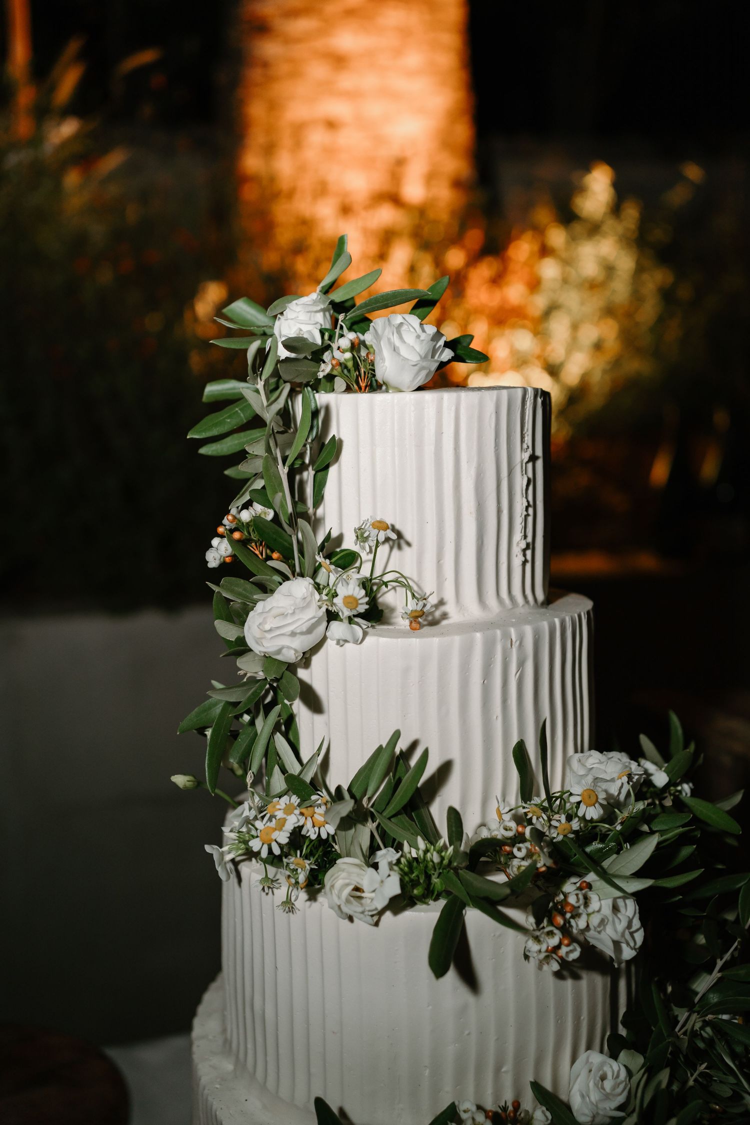 White tiered wedding cake decorated with textured vertical lines and adorned with fresh greenery and white flowers.