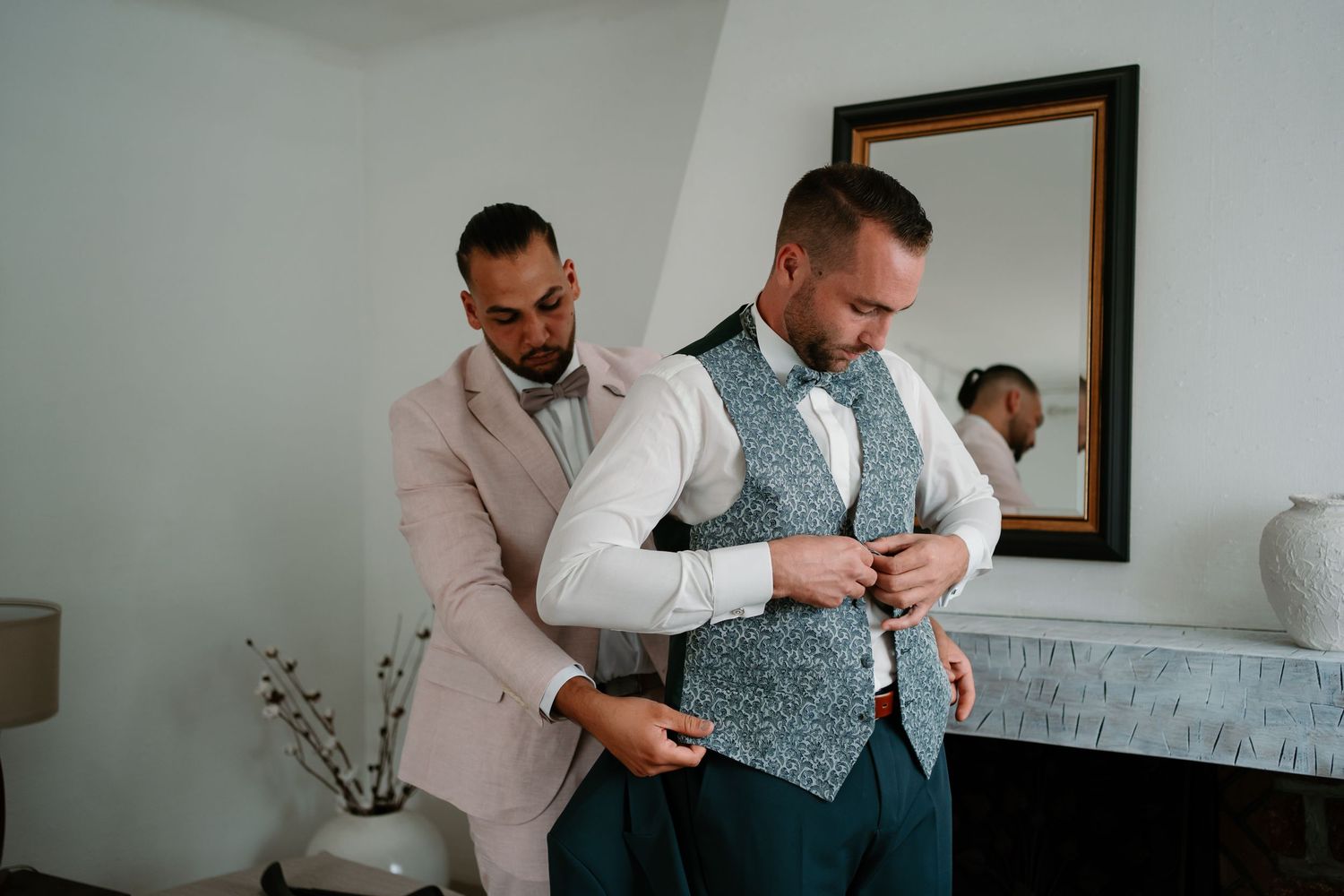 Two people in formal attire help each other get dressed in a room with a mirror and white walls.
