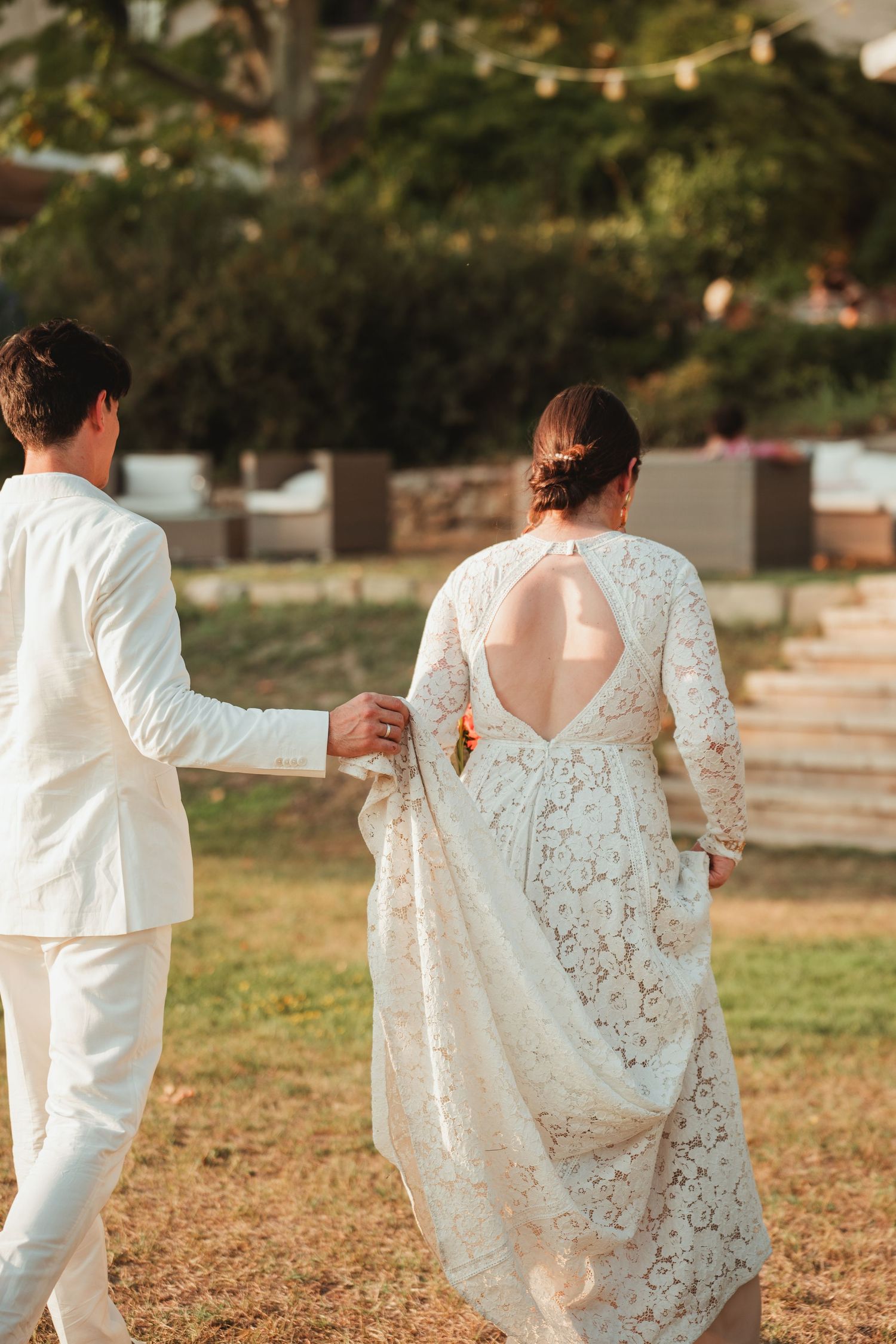 A couple in white wedding attire holding hands and walking through an outdoor garden setting.