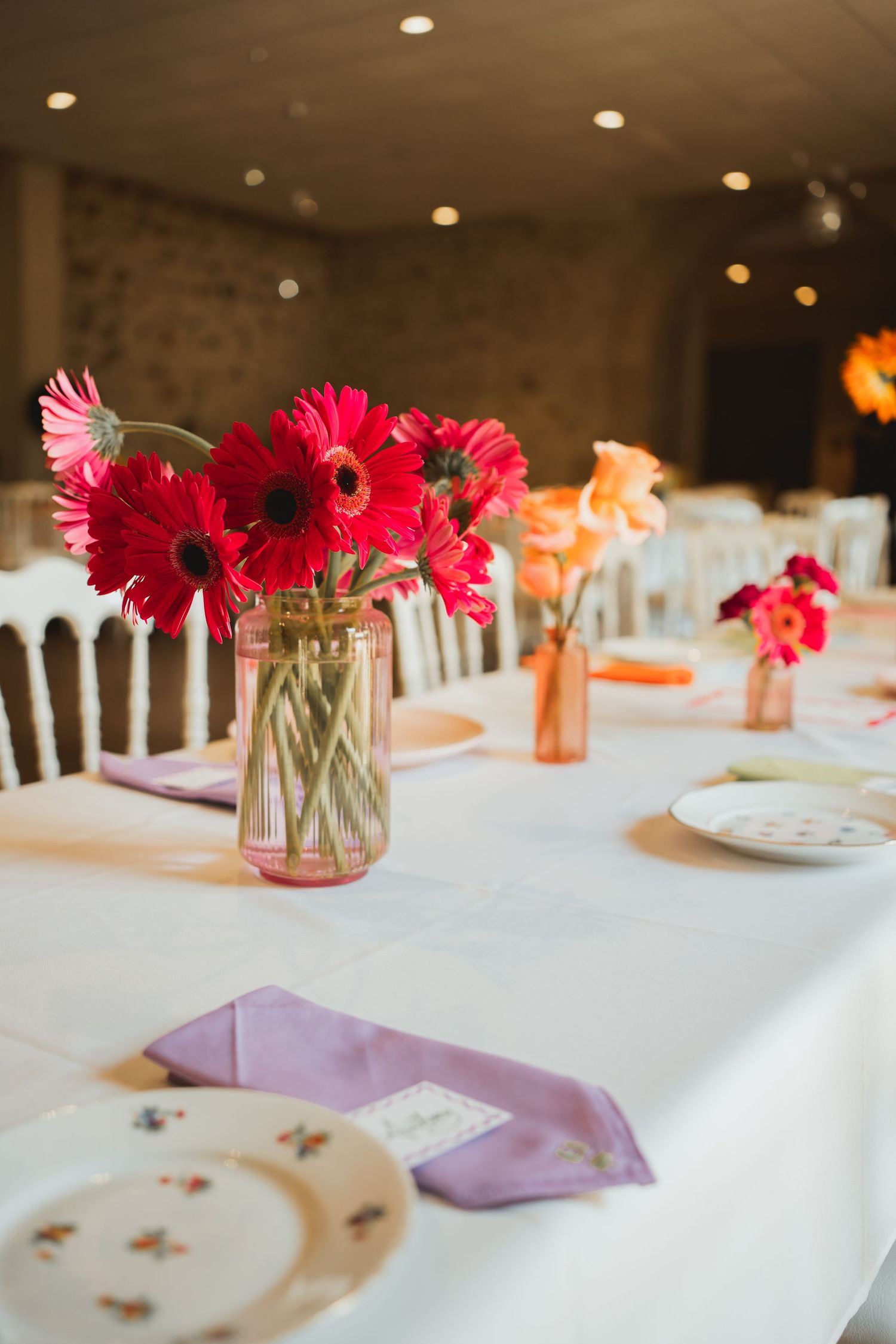 Pink gerbera daisies in mason jars serve as simple yet elegant centerpieces on a white tablecloth at a wedding reception.