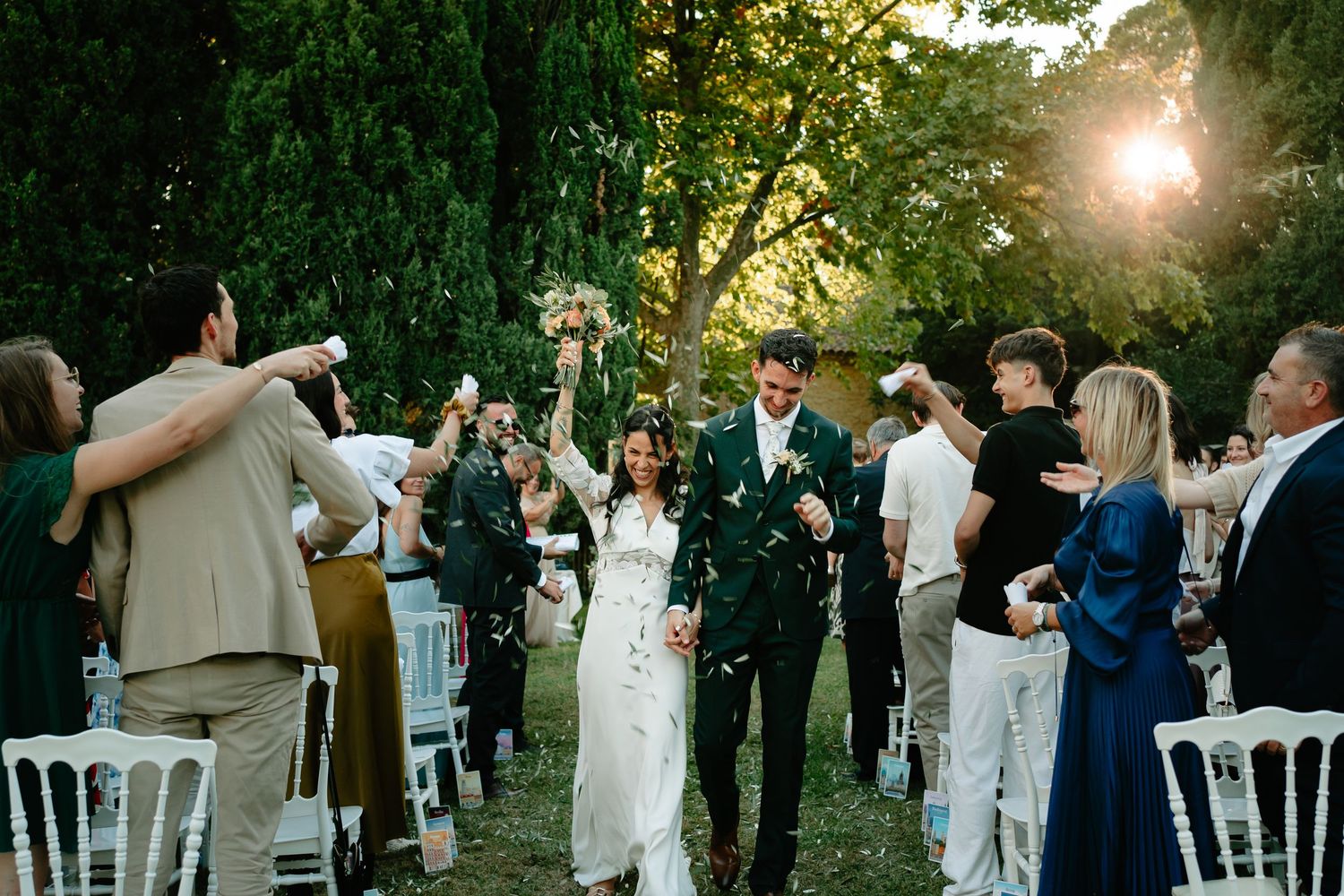 Outdoor wedding ceremony at sunset with guests in white and navy blue attire gathered under trees.