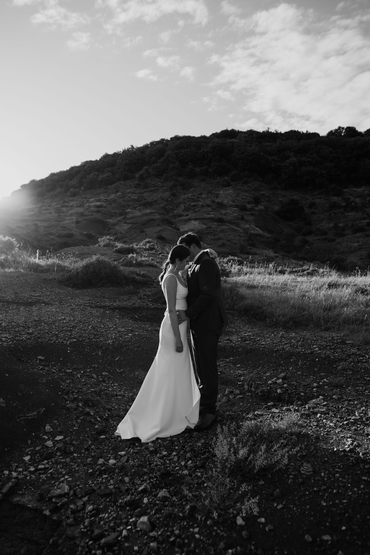 Black and white photo of a bride and groom embracing on rocky terrain with a sunlit hill backdrop.