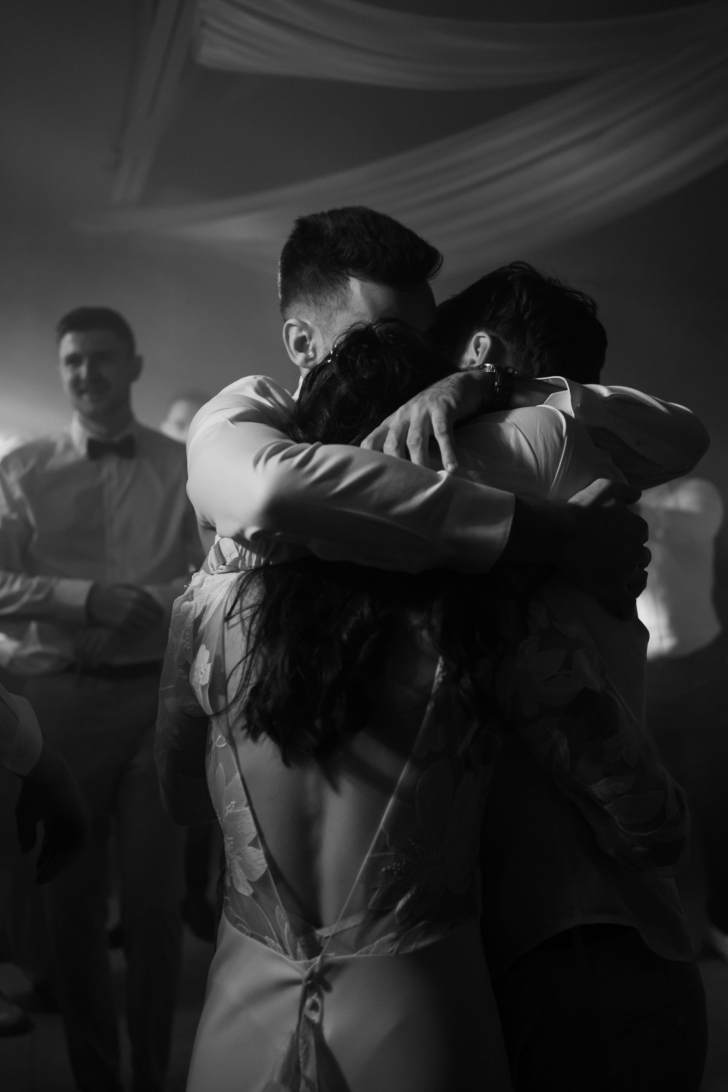 People embrace in an emotional group huddle on a dimly lit dance floor with white draping visible in the background.
