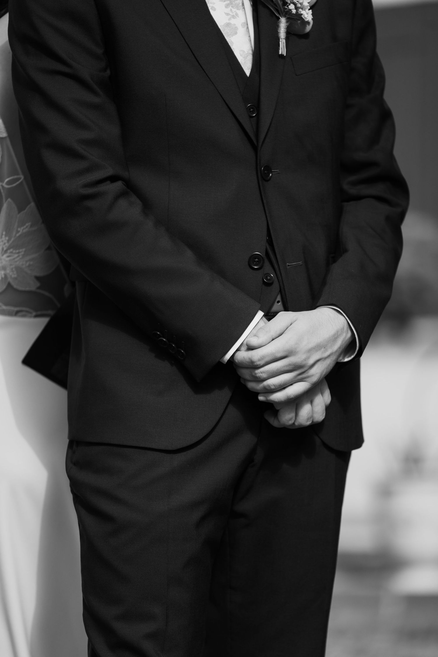 Black and white photo showing groomsmen in formal suits standing with hands clasped in front during wedding ceremony.