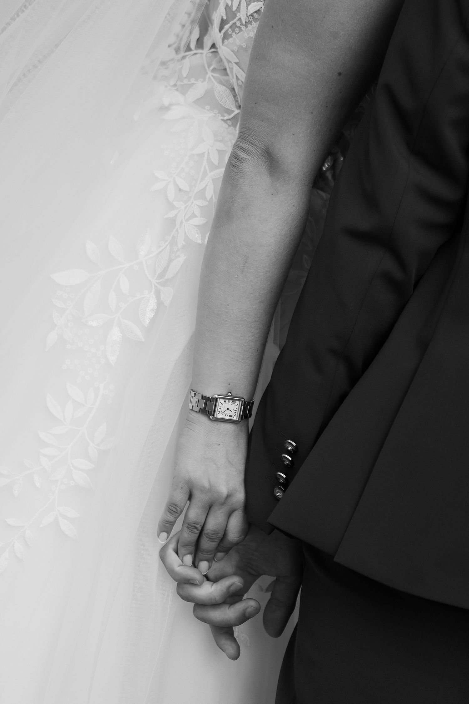 Close-up black and white photo of a bride and groom holding hands, showing wedding rings and formal attire.