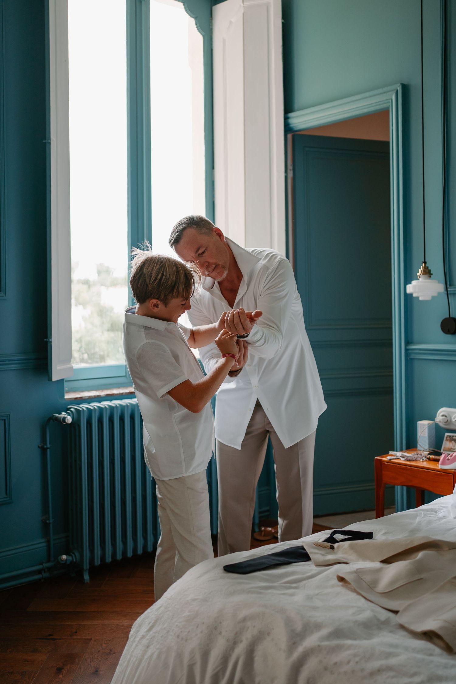 A couple in white outfits shares an intimate moment in a room with teal walls and large windows.