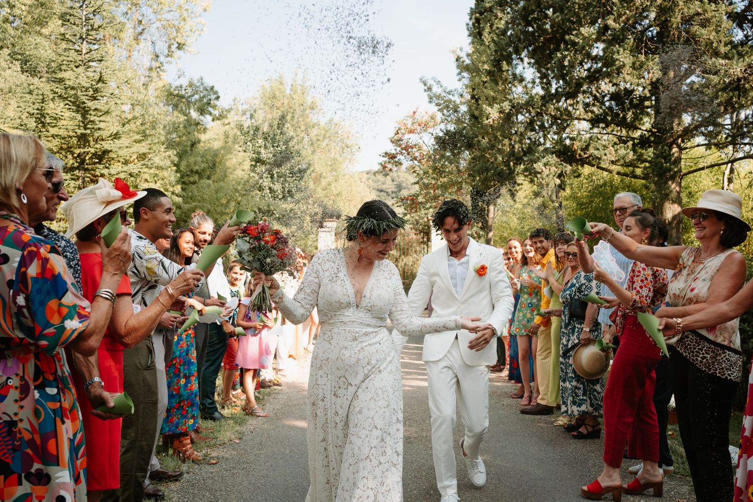 Wedding couple walks through joyful crowd throwing confetti on tree-lined path during outdoor celebration.