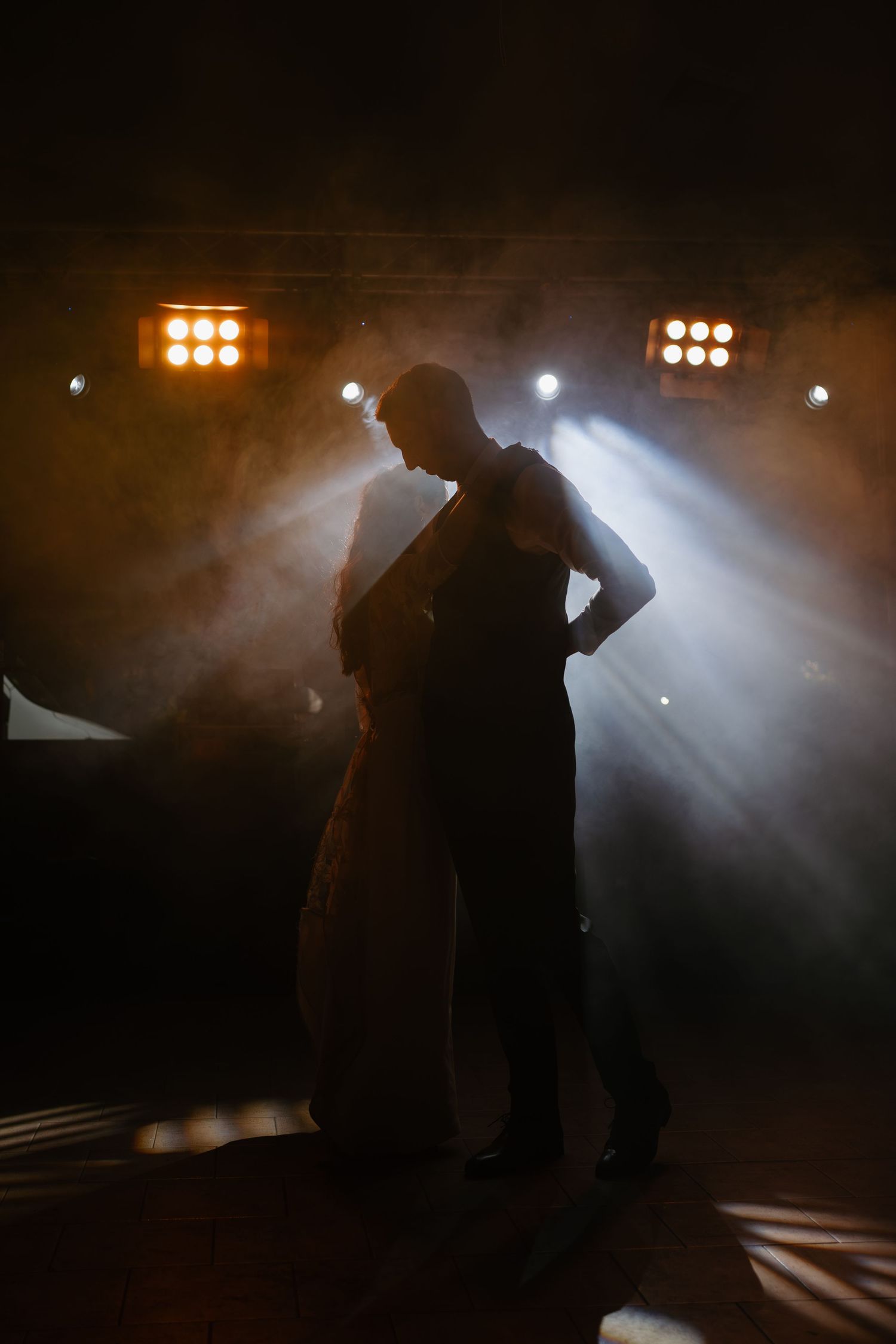 Silhouetted figures perform on a smoky stage with dramatic backlighting and spotlights creating an atmospheric scene.