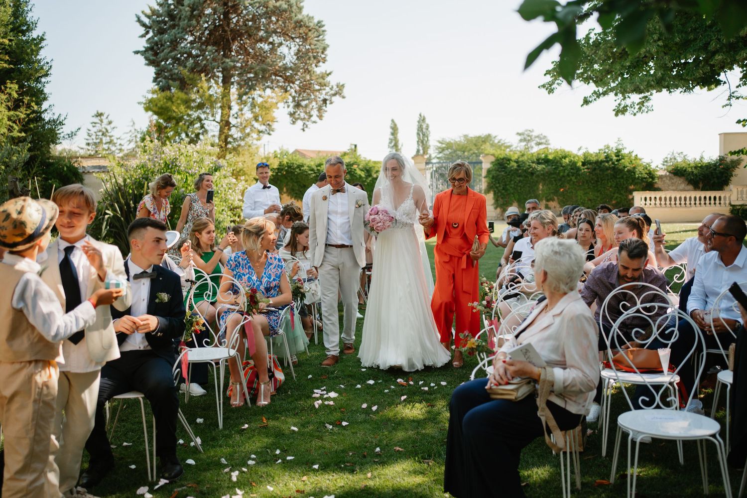Outdoor garden wedding ceremony with guests seated on white chairs surrounded by lush greenery and scattered flower petals.