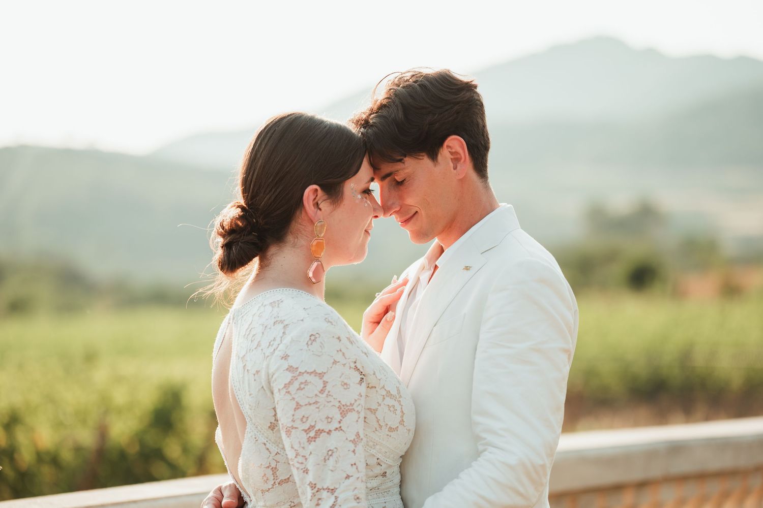 A couple in wedding attire embraces in a sunlit vineyard with rolling hills in the background.
