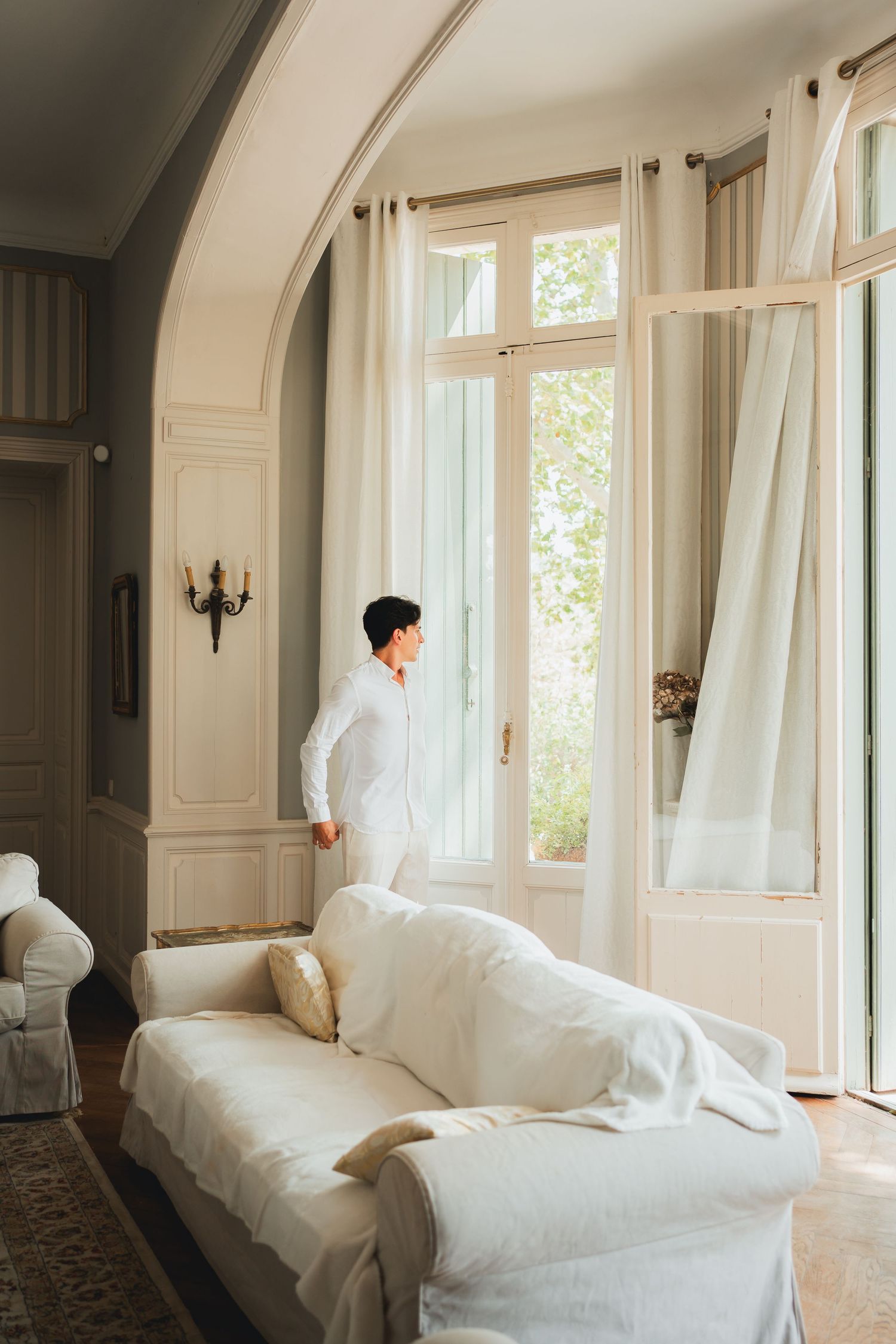 Bright white bedroom with curved walls and large windows features flowing sheer curtains and unmade bedding in natural light.