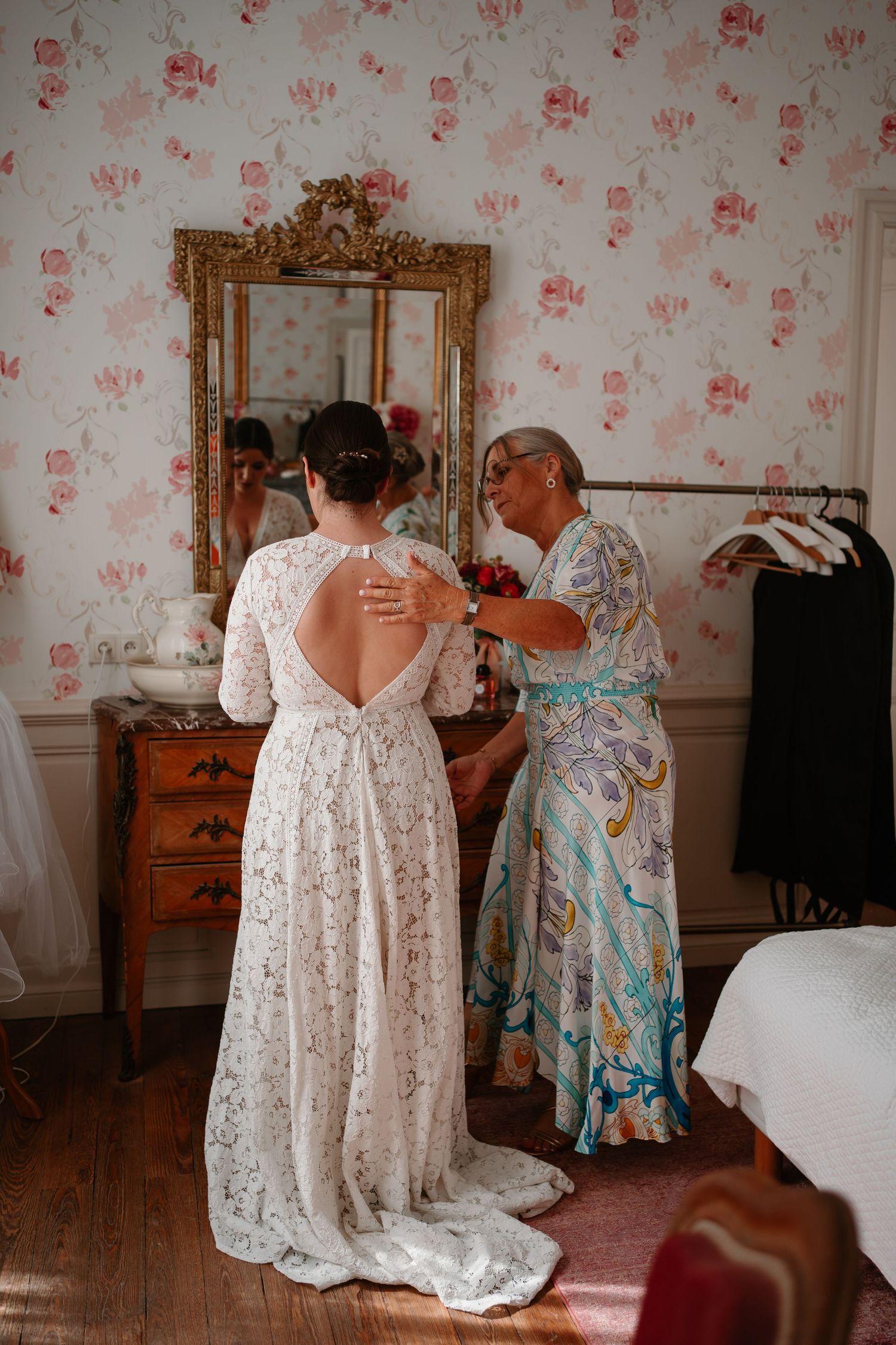 A white lace wedding dress being adjusted in front of an ornate gold mirror with floral wallpaper in the background.
