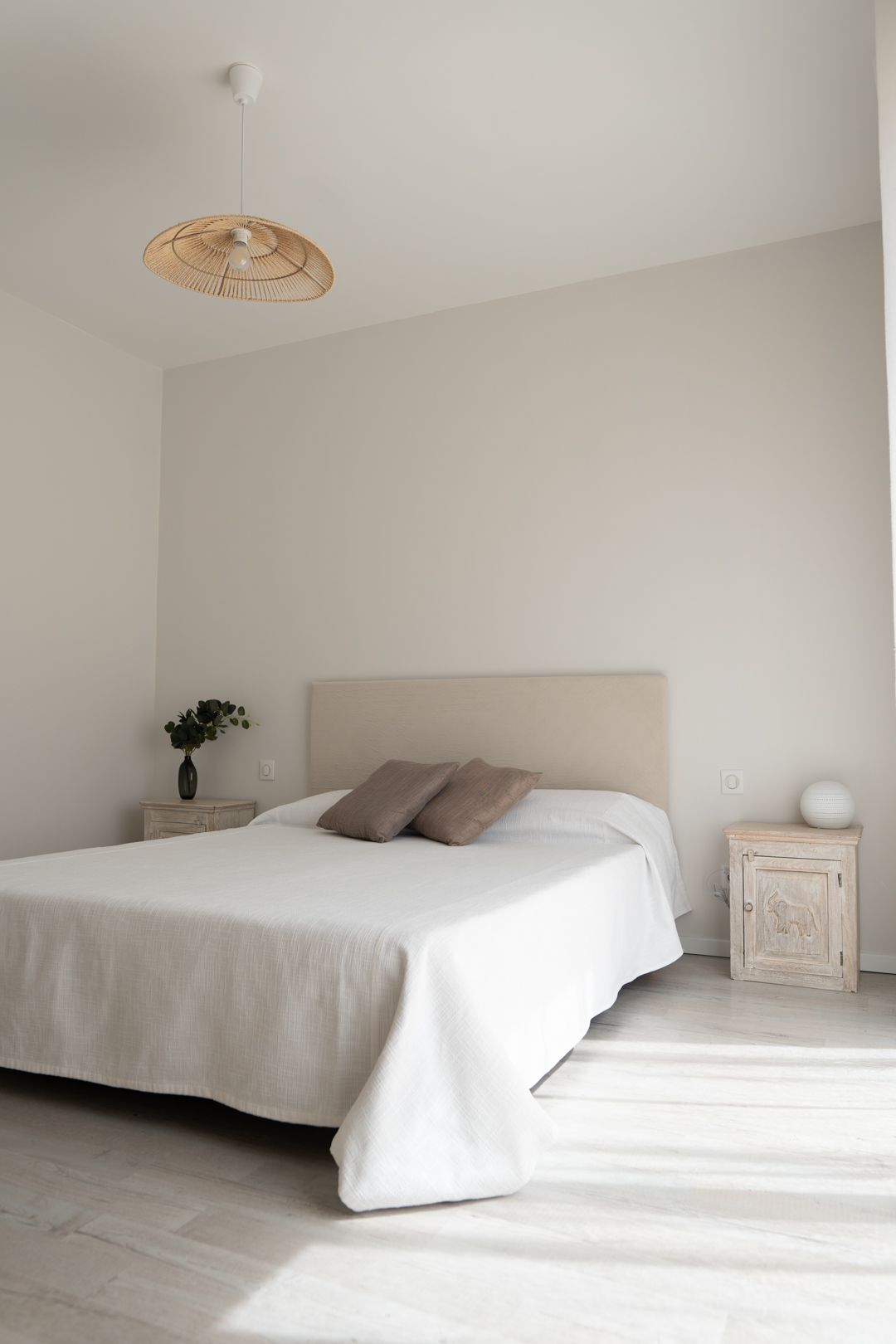 A minimalist white bedroom with a simple bed, light wooden floors, and natural lighting streaming through the windows.