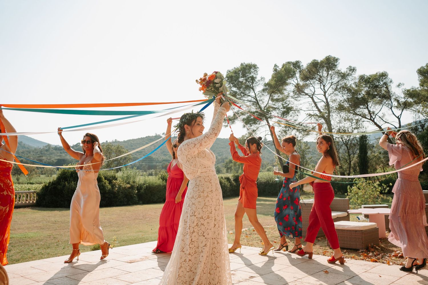 Wedding party dances in a line with arms raised under a white canopy at an outdoor celebration.