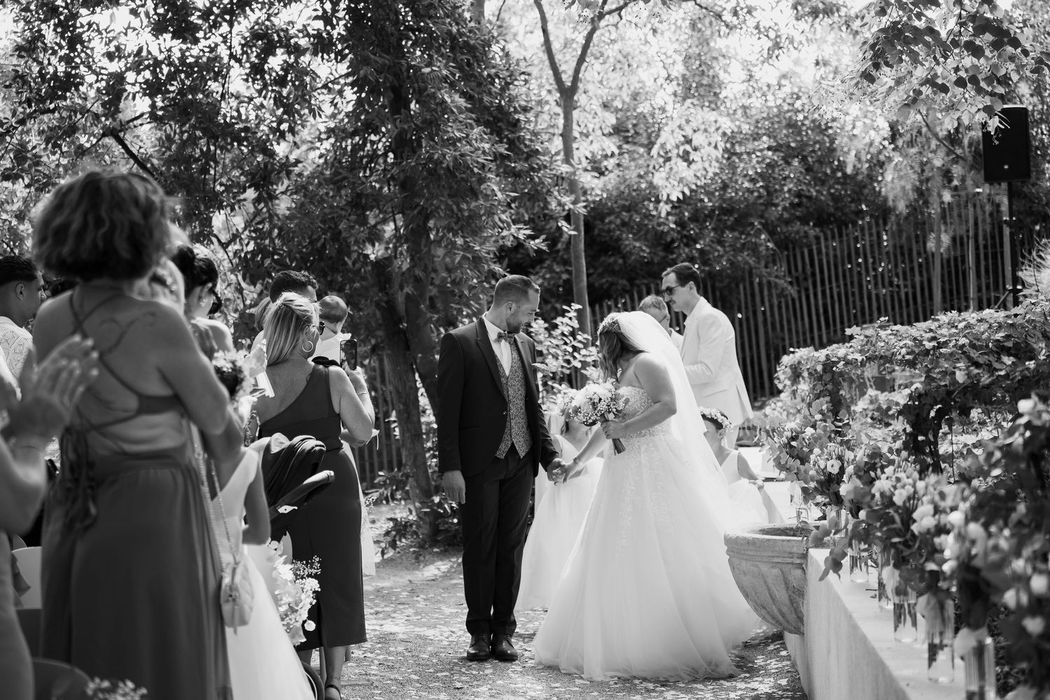 Black and white outdoor wedding ceremony in a garden with guests gathered along a floral-lined path.