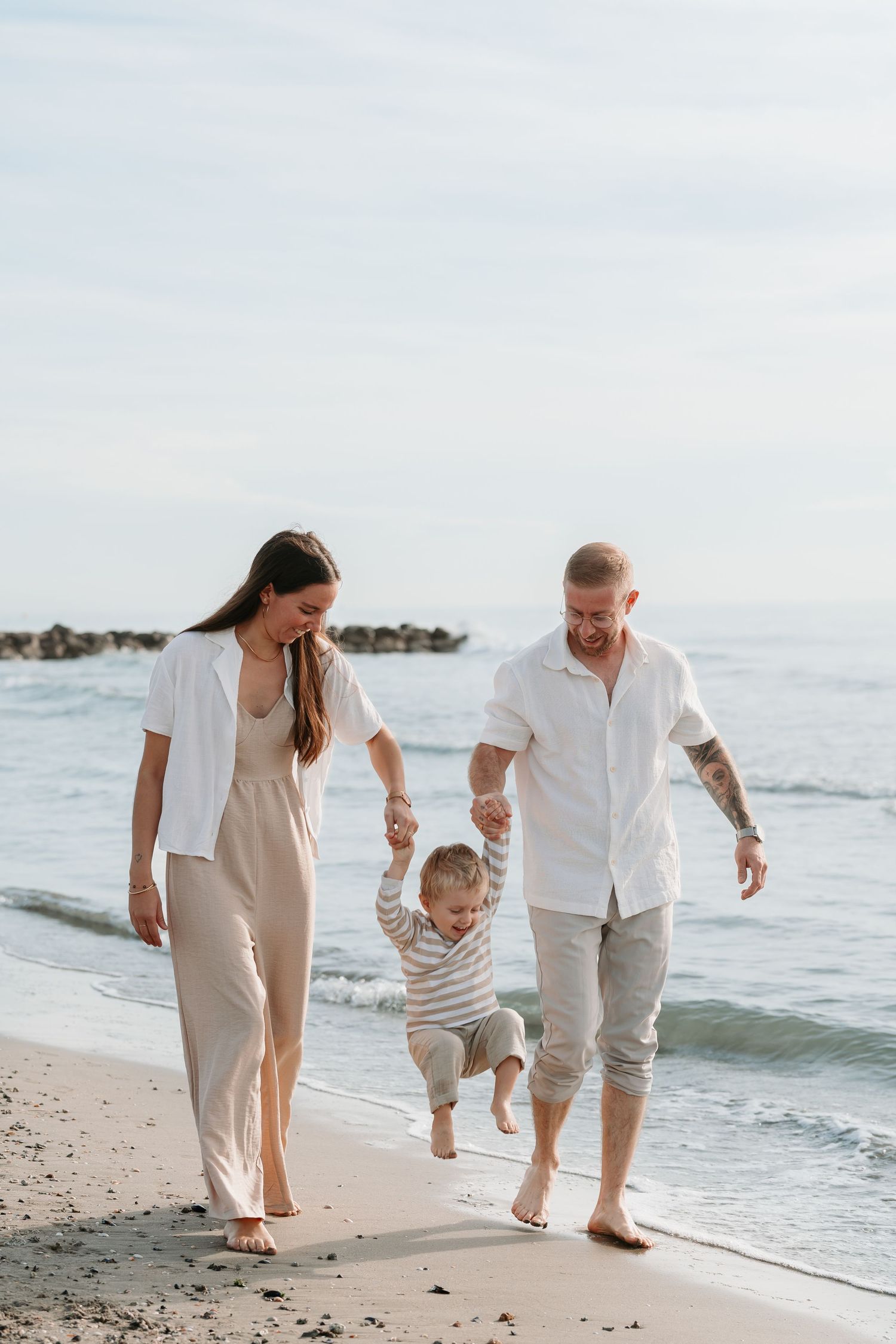 Family in coordinated white and beige outfits walking together along a sunny beach shoreline.