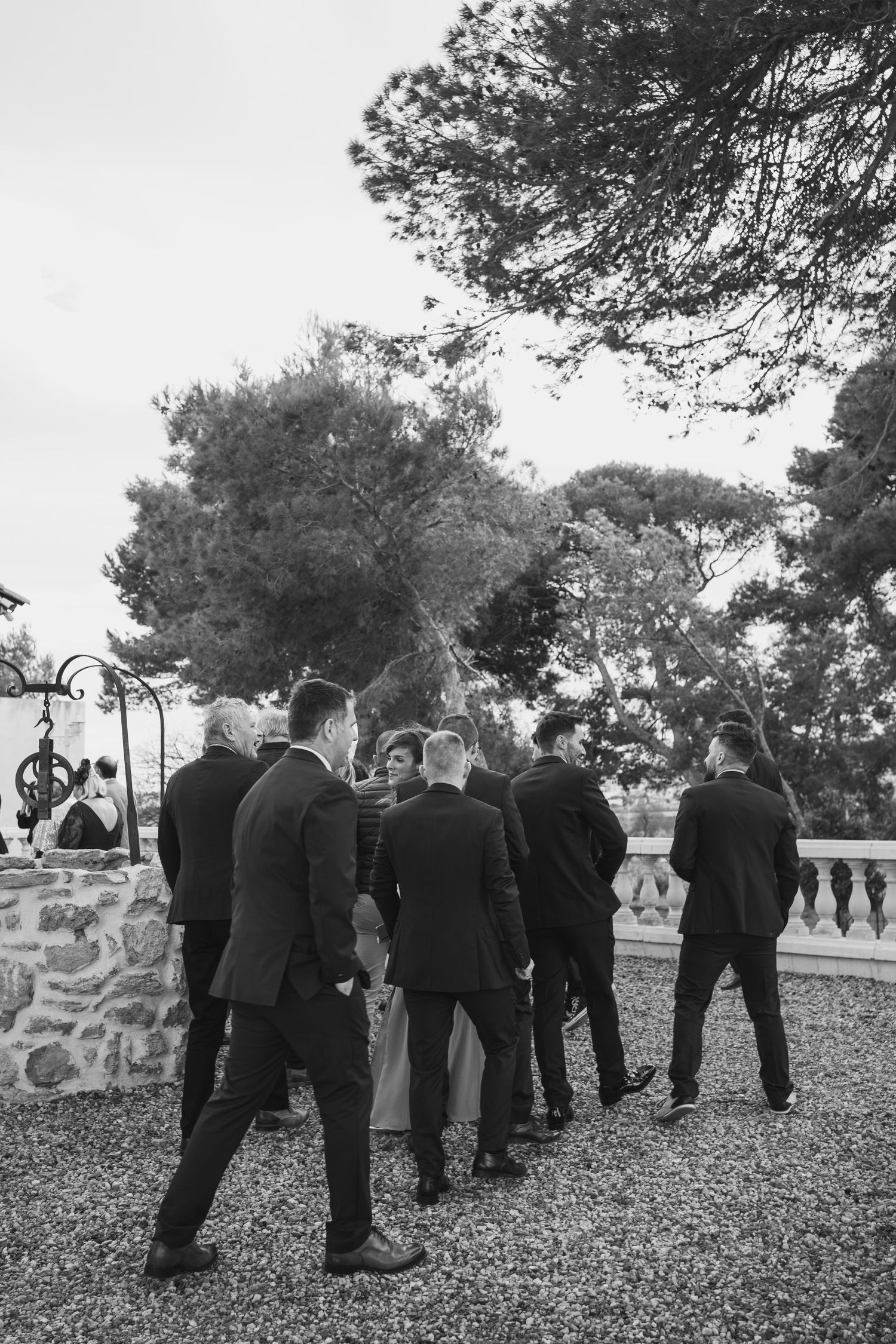 Groomsmen in dark suits gather for formal group photo on gravel path.