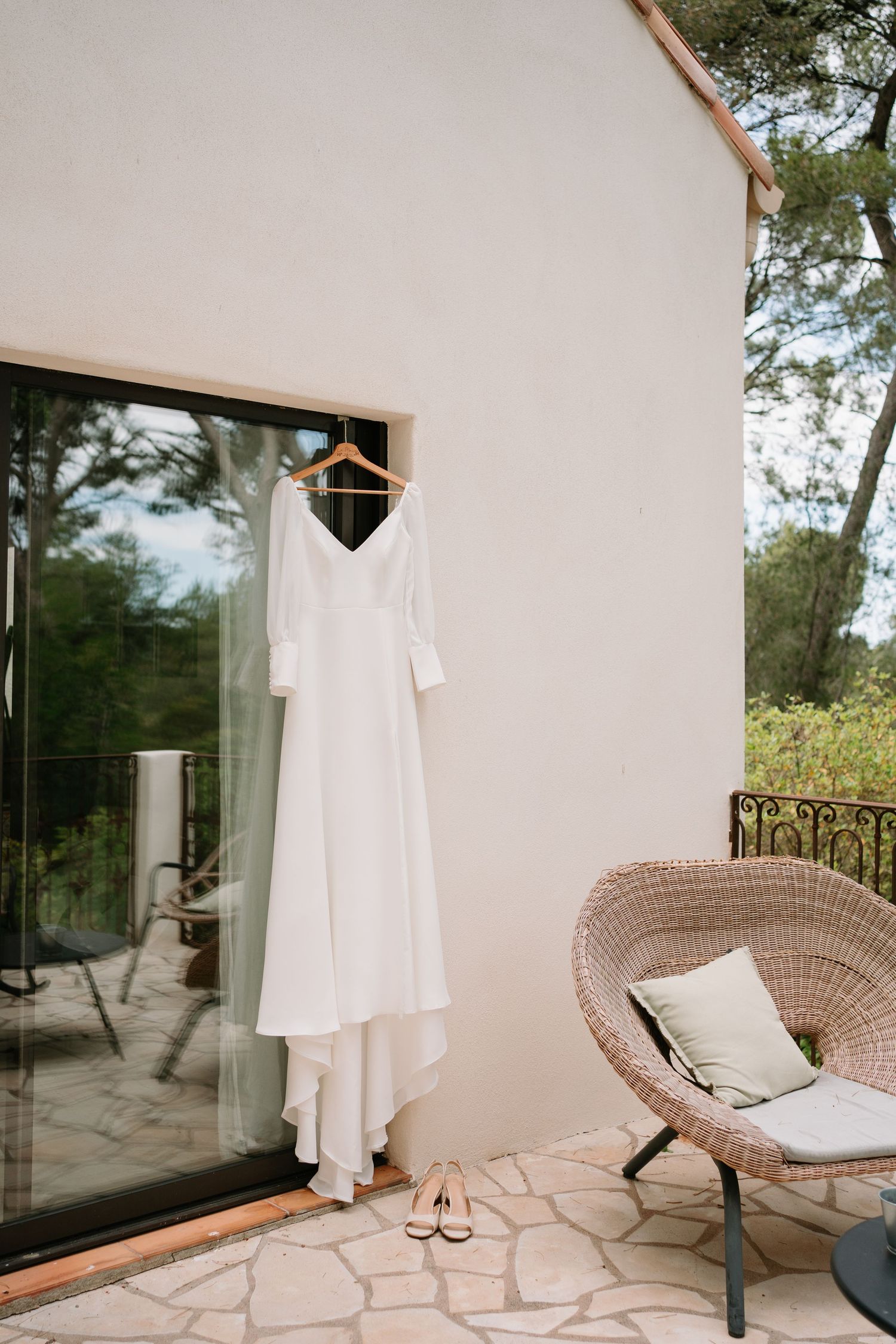 Elegant white long-sleeve wedding dress hanging on wooden hanger outside villa door with wicker chair and bridal shoes.