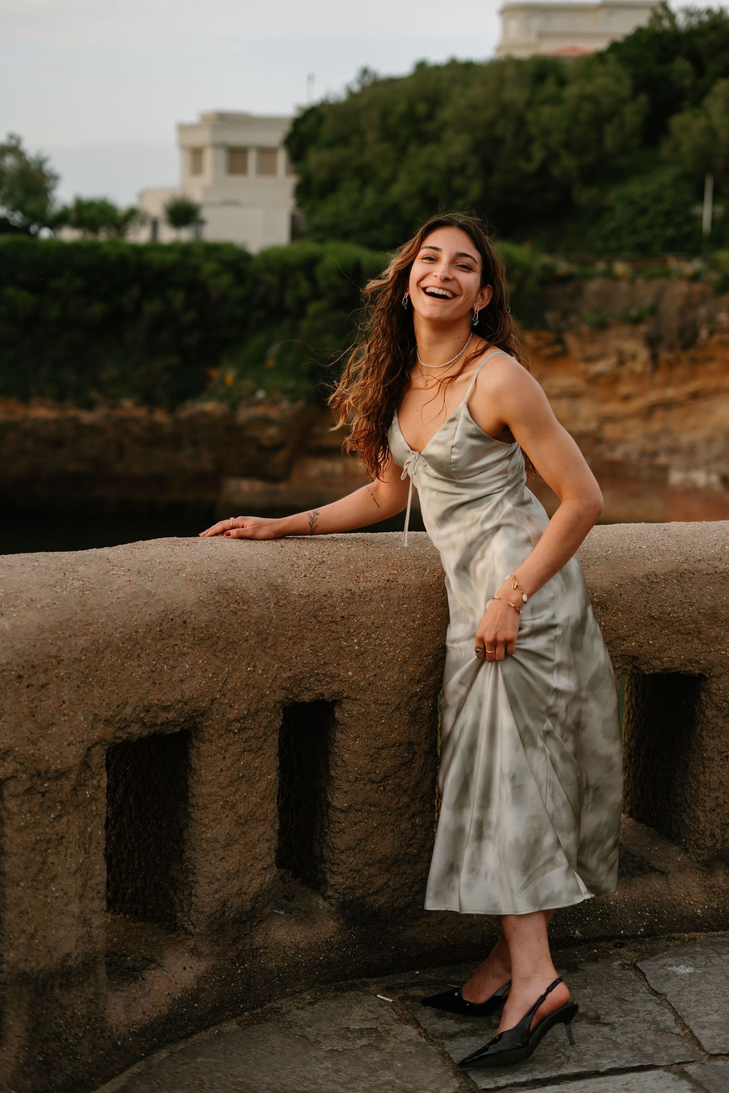 Woman in silver dress leans against stone balustrade with coastal view.