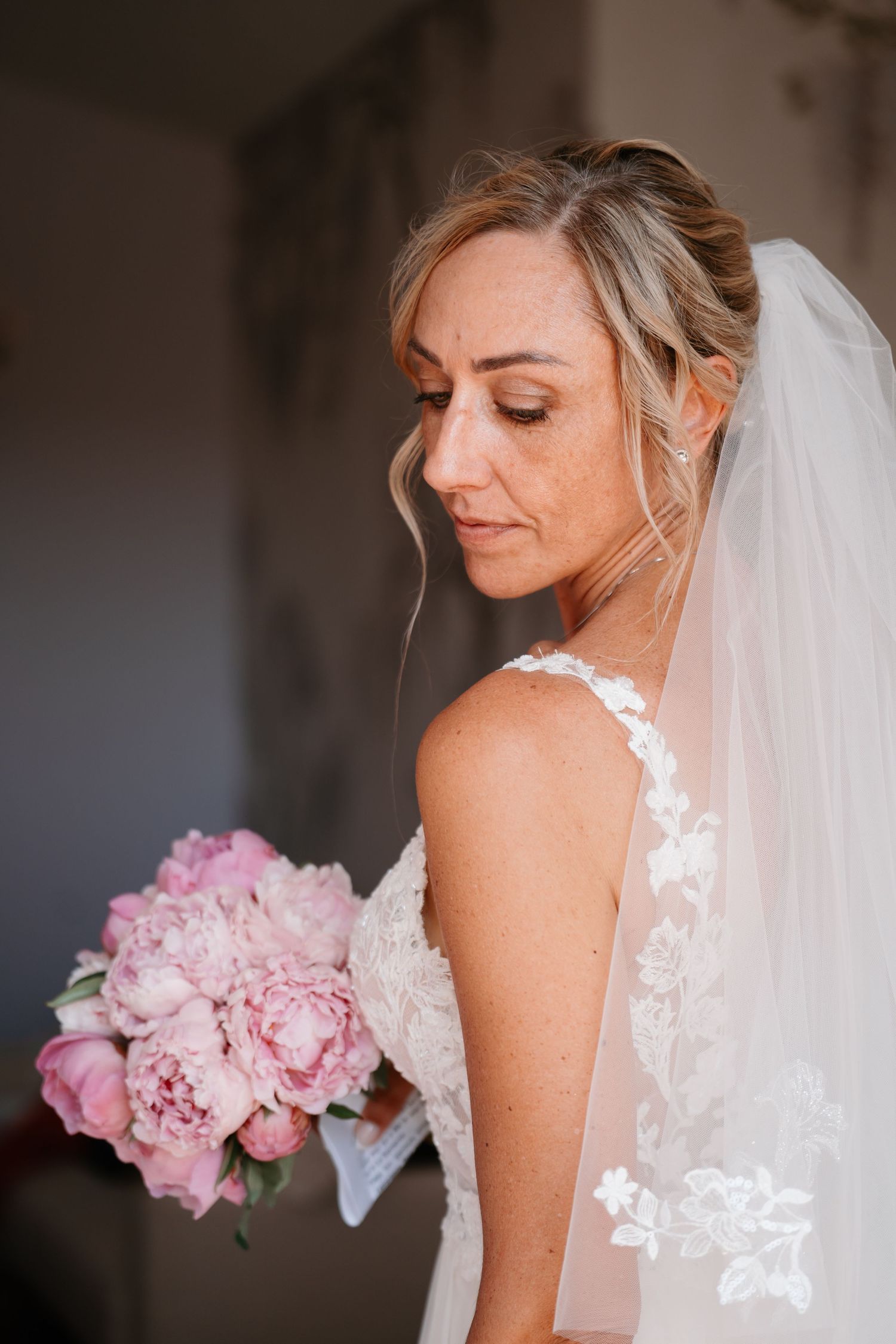 A bride in a white lace wedding dress with a veil holds a bouquet of pink peonies against a soft blurred background.