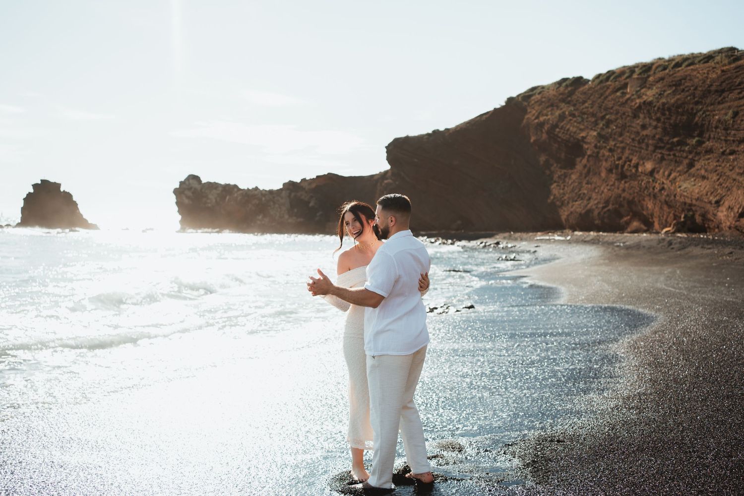 Couple in white outfits sharing romantic moments on a scenic beach with rocky cliffs at sunset.
