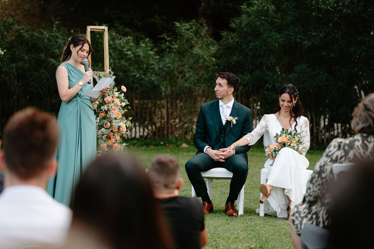 A wedding ceremony takes place outdoors with attendees gathered in a garden setting during a sunny day.