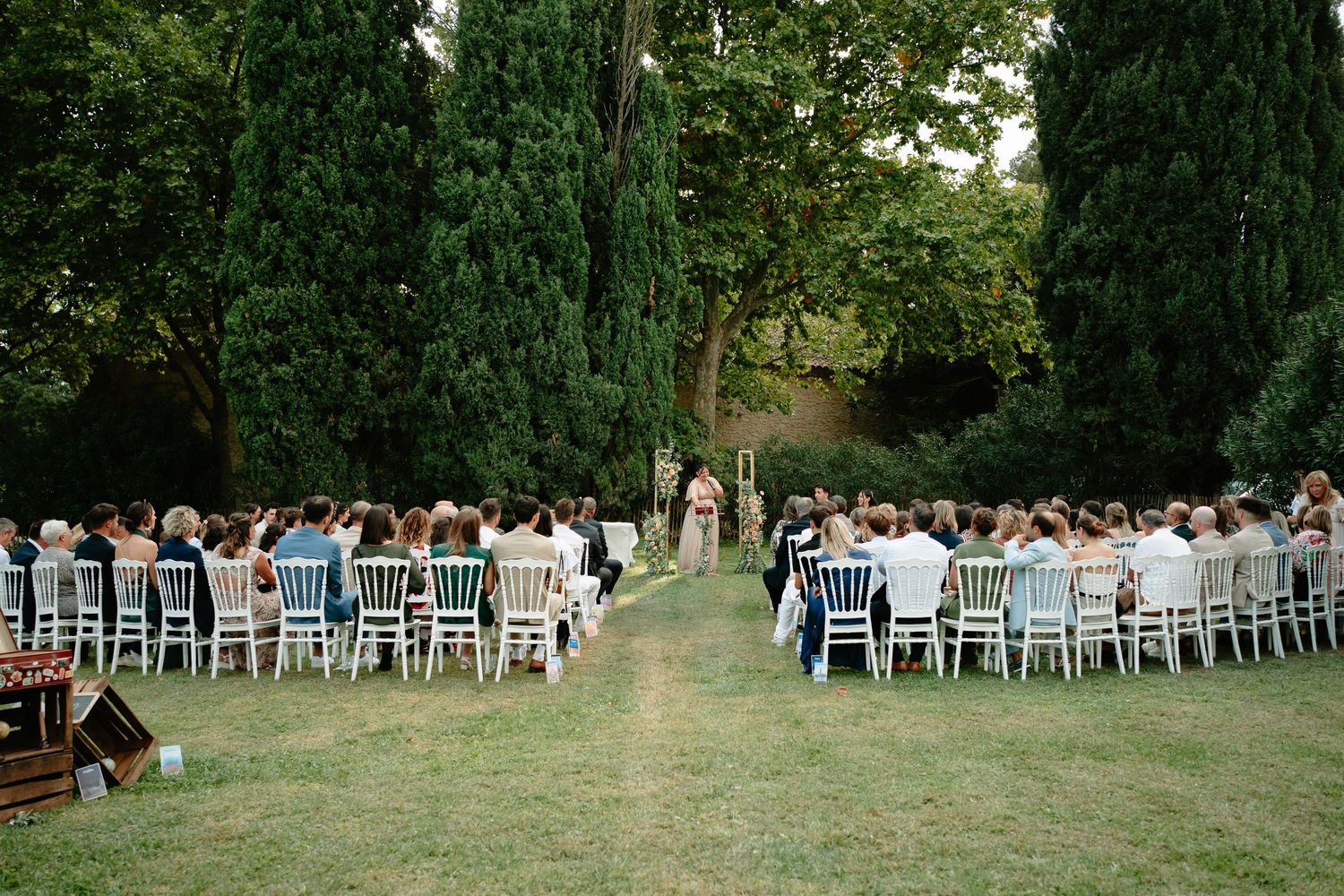 Outdoor wedding ceremony on a green lawn with white chairs, guests seated, and tall trees in a garden setting.