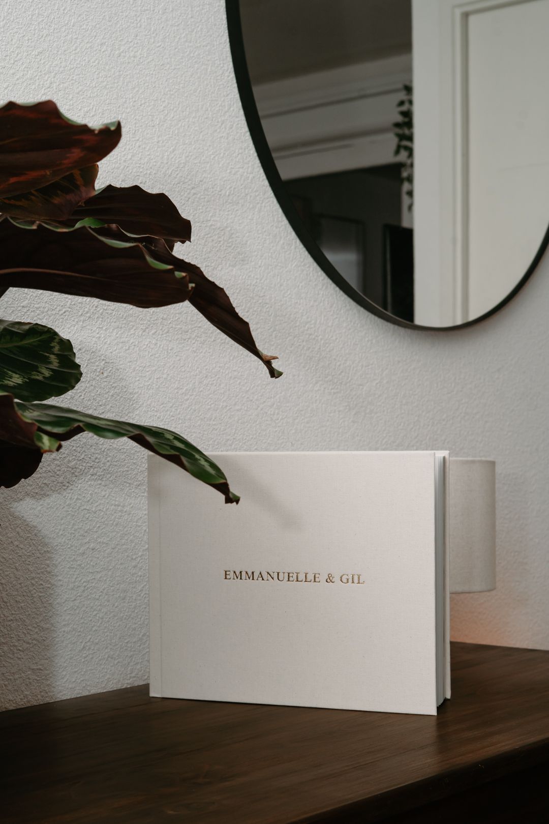 A minimalist white photo album with text 'EMMANUELLE & GIL' sits on a dark wooden surface near a round mirror.