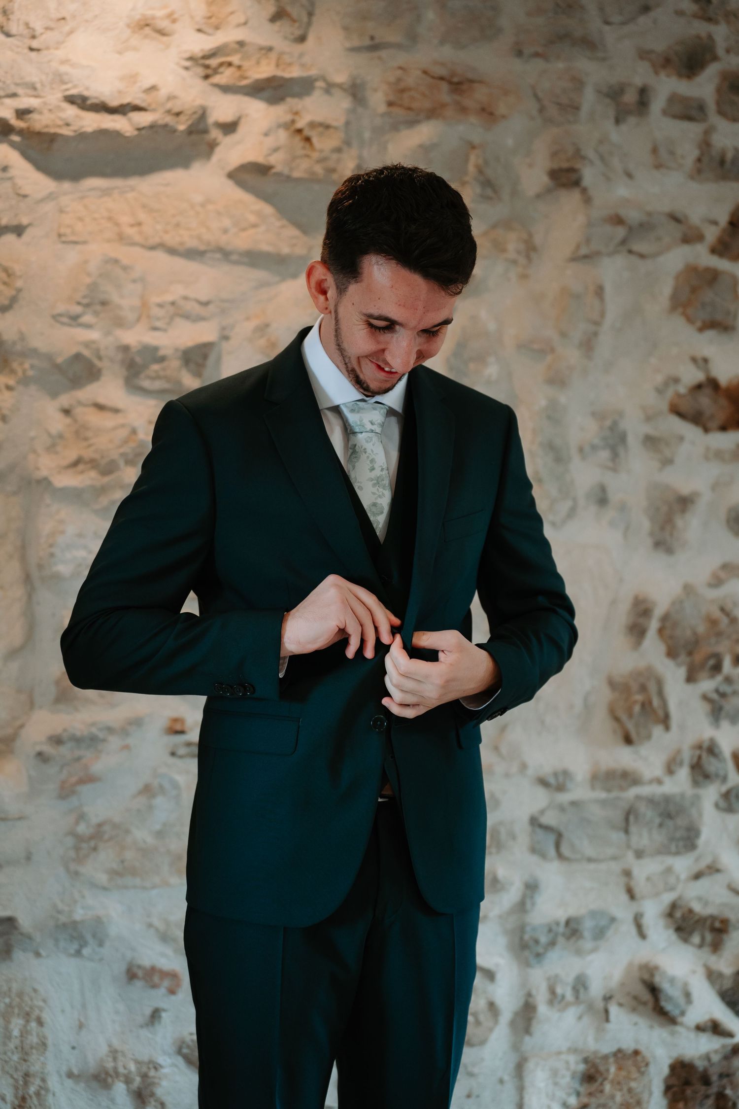 Man in a black suit adjusts his tie while standing against a rustic stone wall.