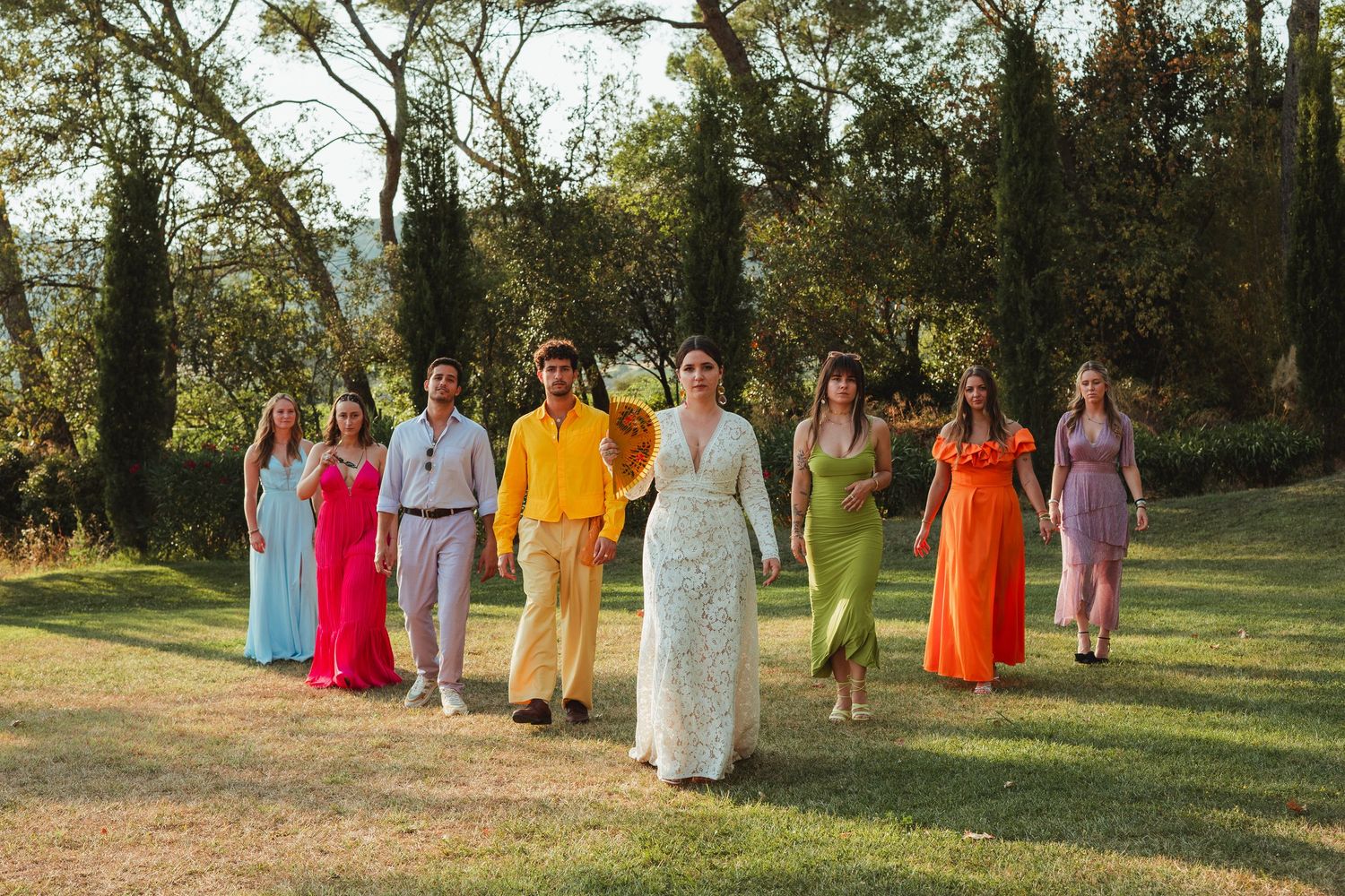A group in colorful formal attire poses together outdoors in a sunlit garden setting.